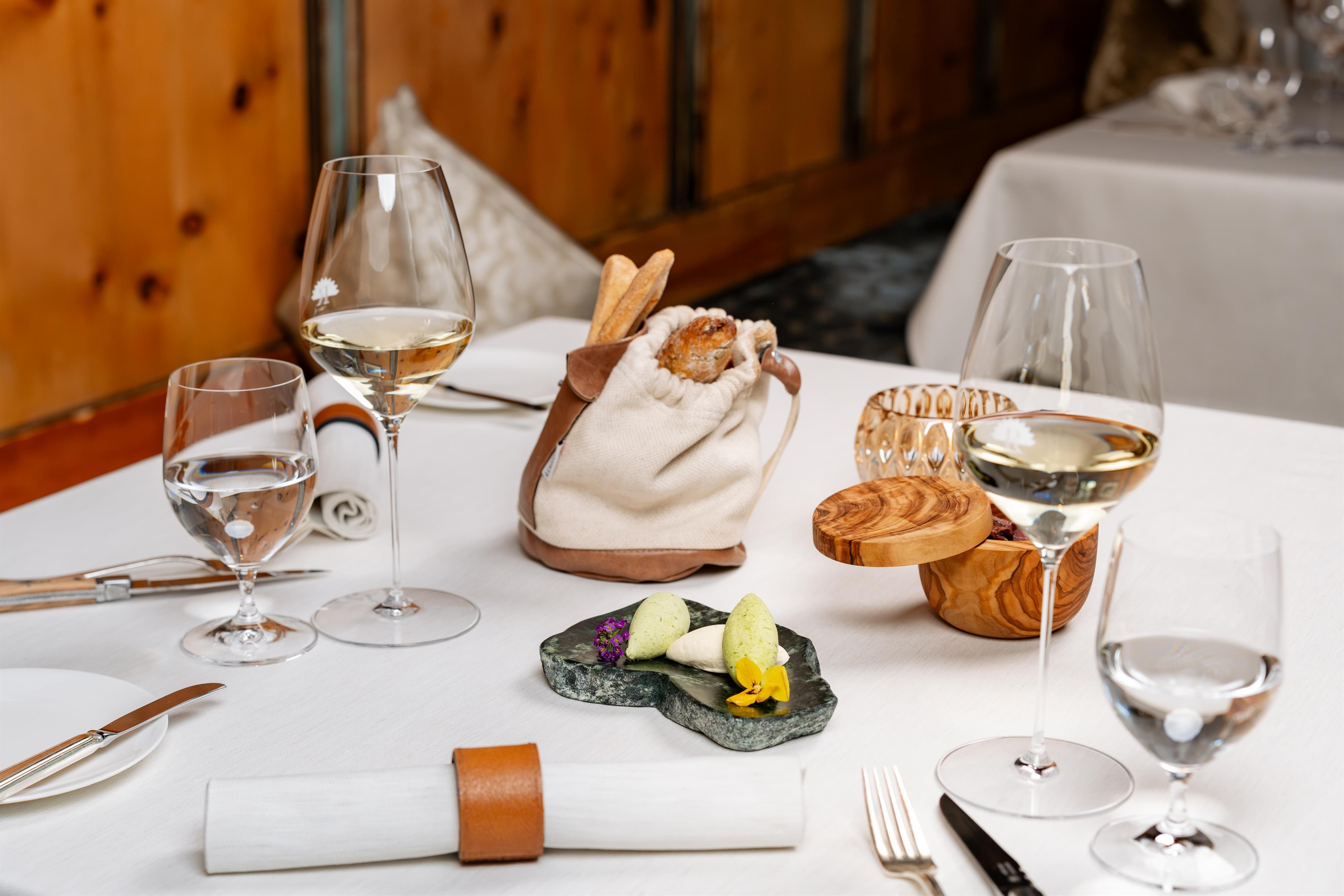 An elegant table with several wine glasses and exquisite dishes. In the center is a small dish on a stone platter, surrounded by stylish cutlery.