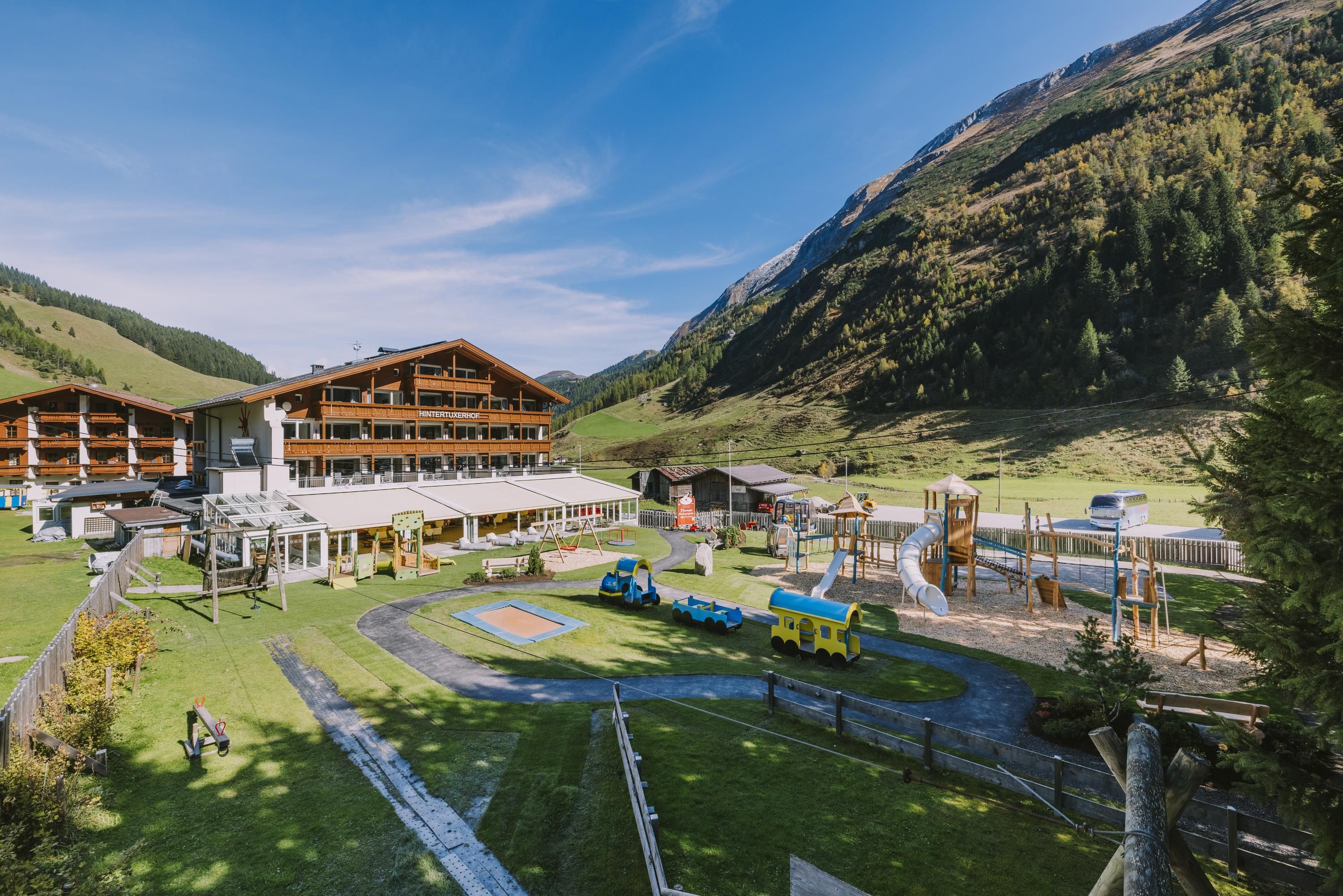 A picturesque landscape with a large hotel building and a playground. In the background, green mountains and a clear sky can be seen.