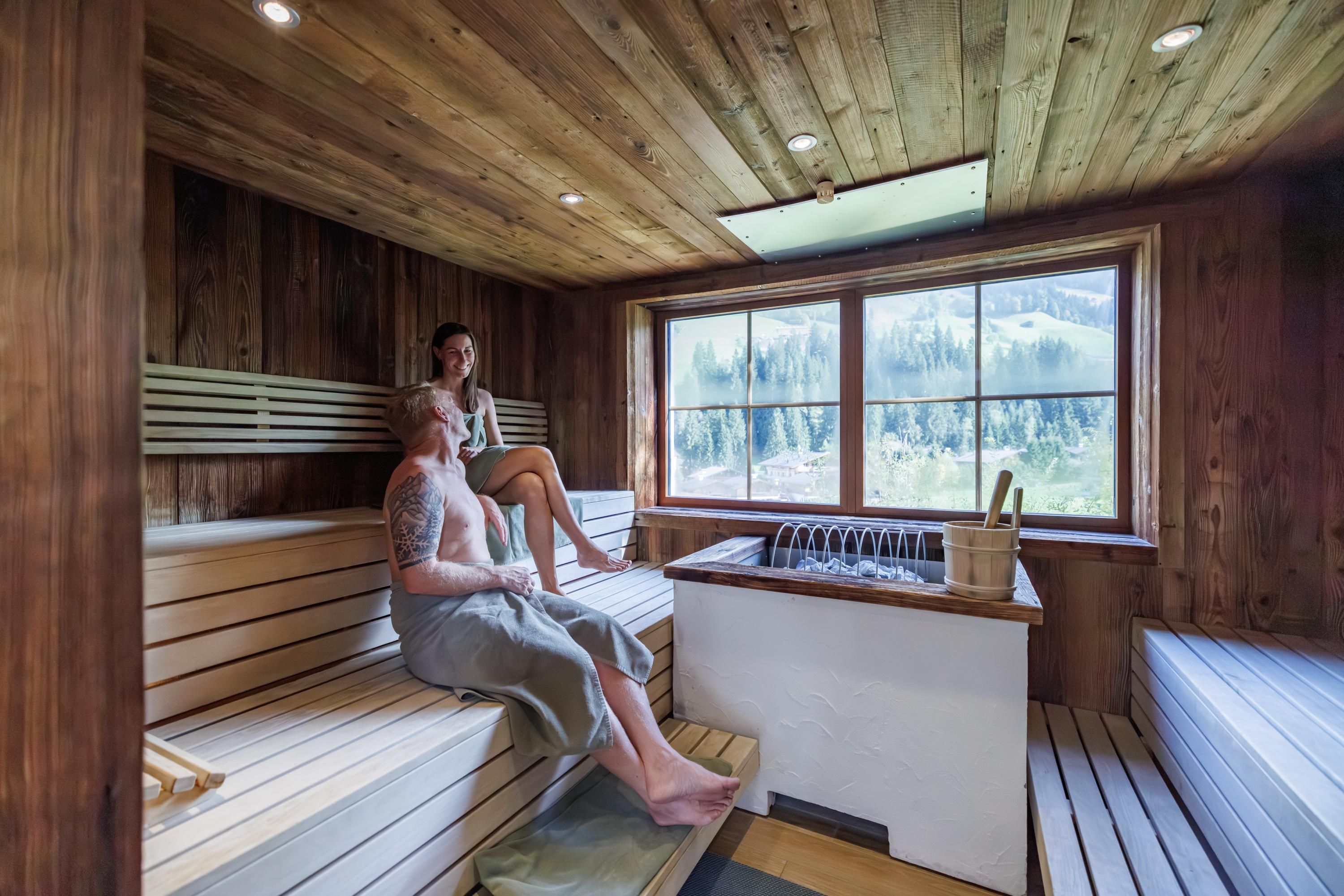 A modern sauna with wooden paneling and large windows. Two women are relaxing comfortably on the benches.