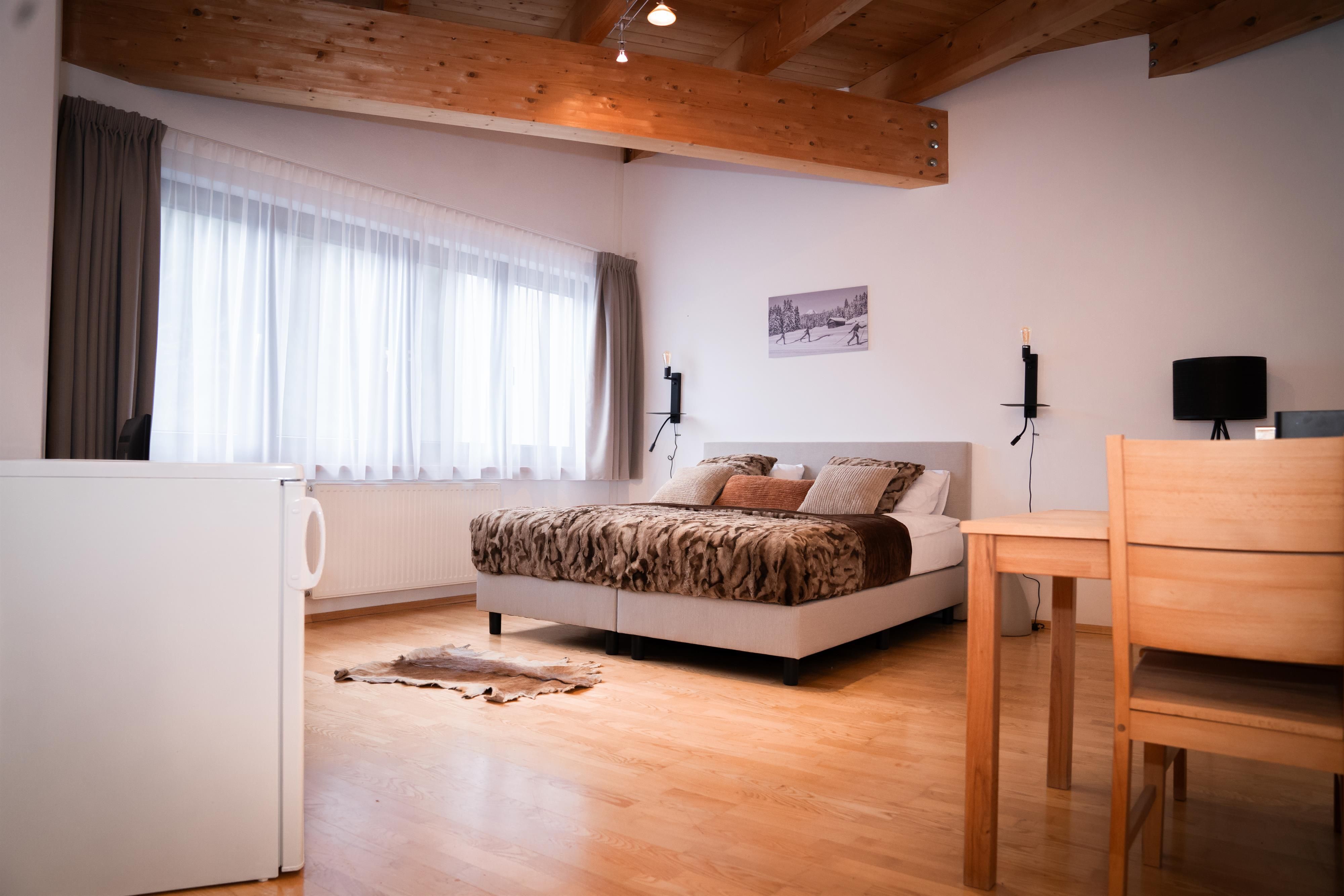 A modern bedroom with a large bed and wooden beams on the ceiling. Next to the bed, there is a table and a refrigerator.