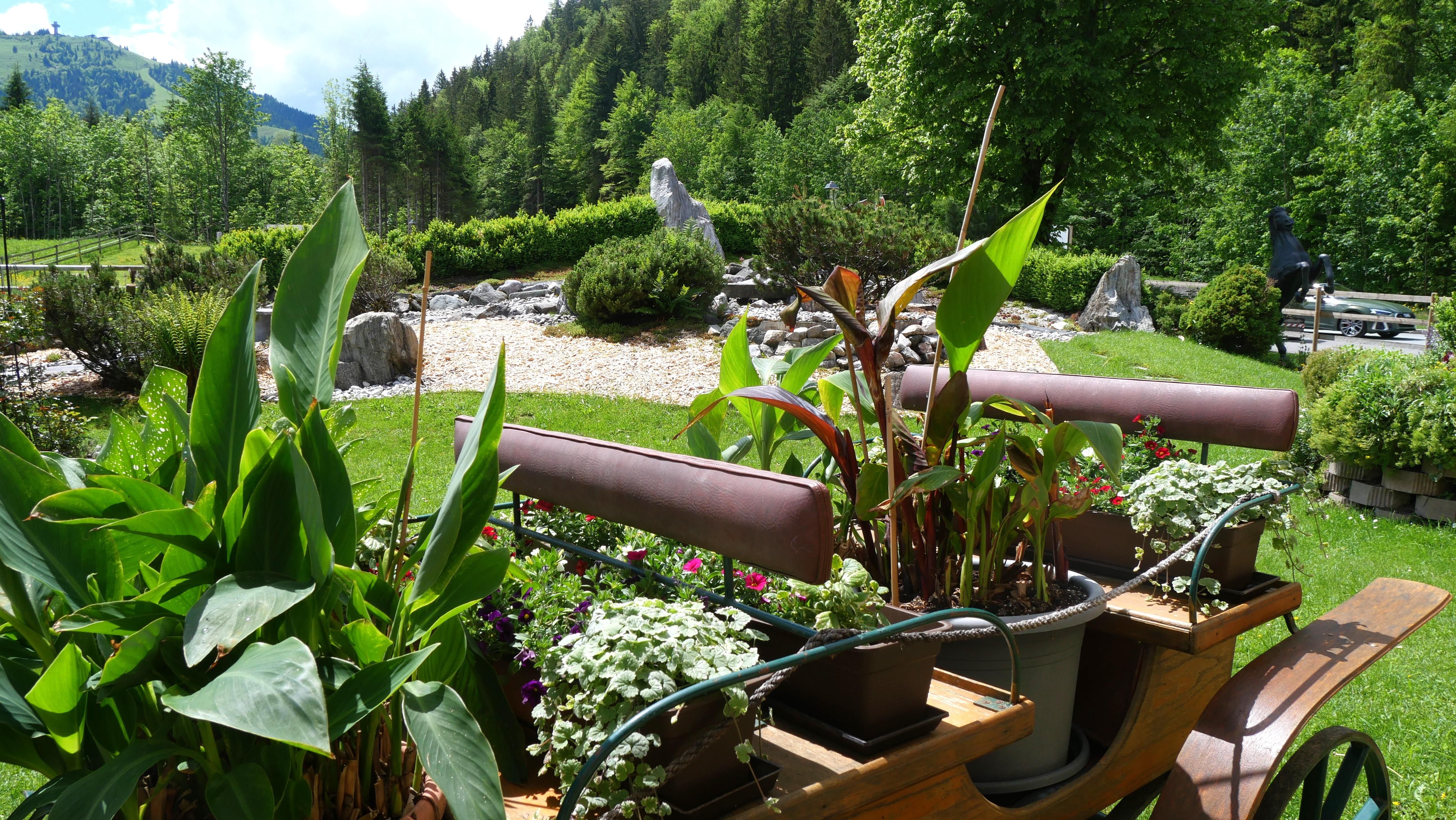 A stylish garden landscape with a wooden structure decorated with flowers and plants. In the background, green trees and a small sculpture garden can be seen.