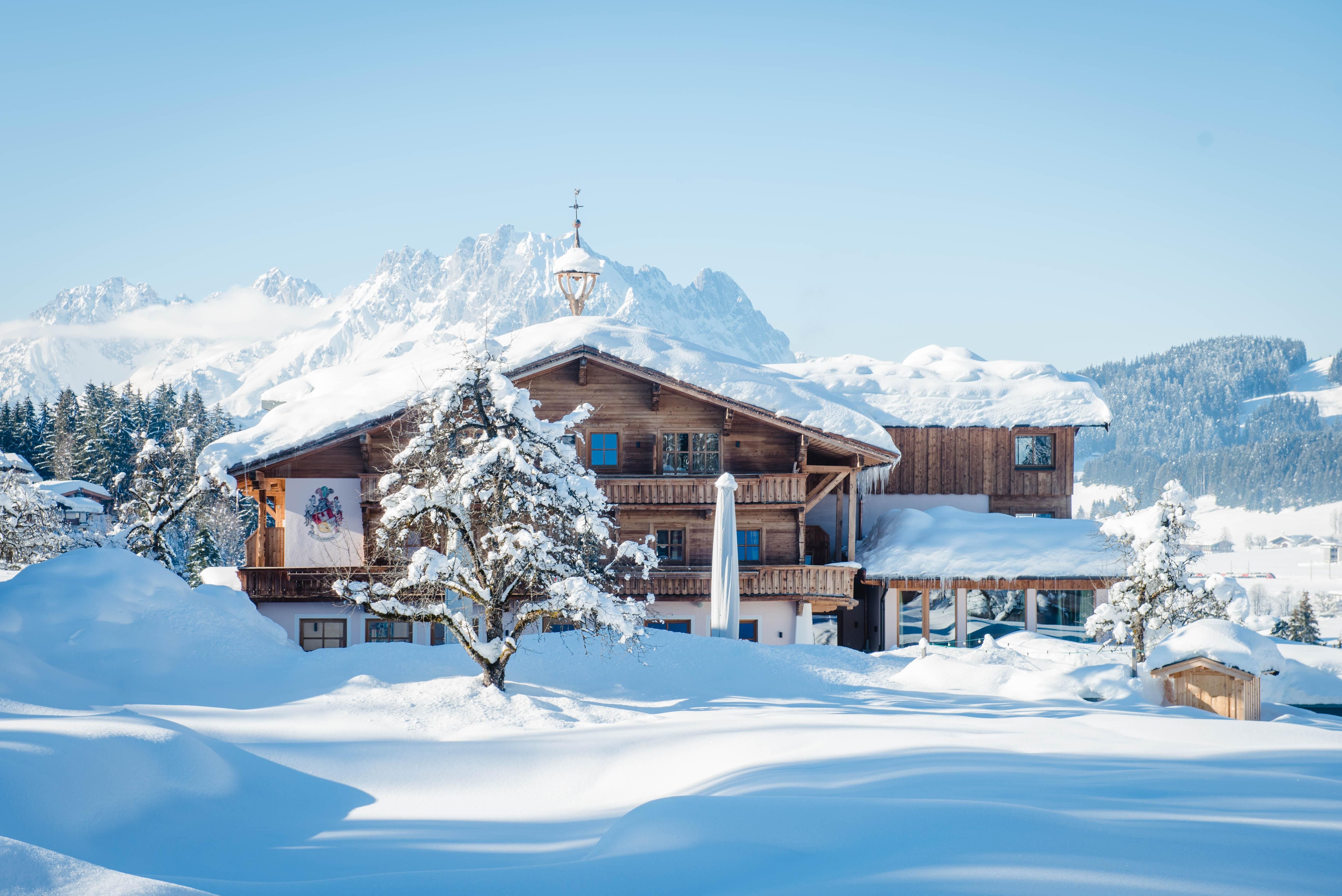 A charming wooden house in the snow, surrounded by snow-covered mountains. The clear blue sky creates a picturesque winter landscape.