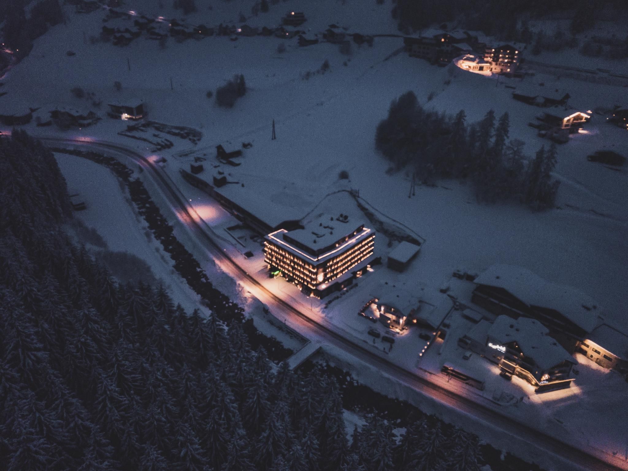 A snow-covered landscape with a illuminated hotel and several small buildings. The streets are slightly icy, and the surroundings exude a calm, wintry atmosphere.