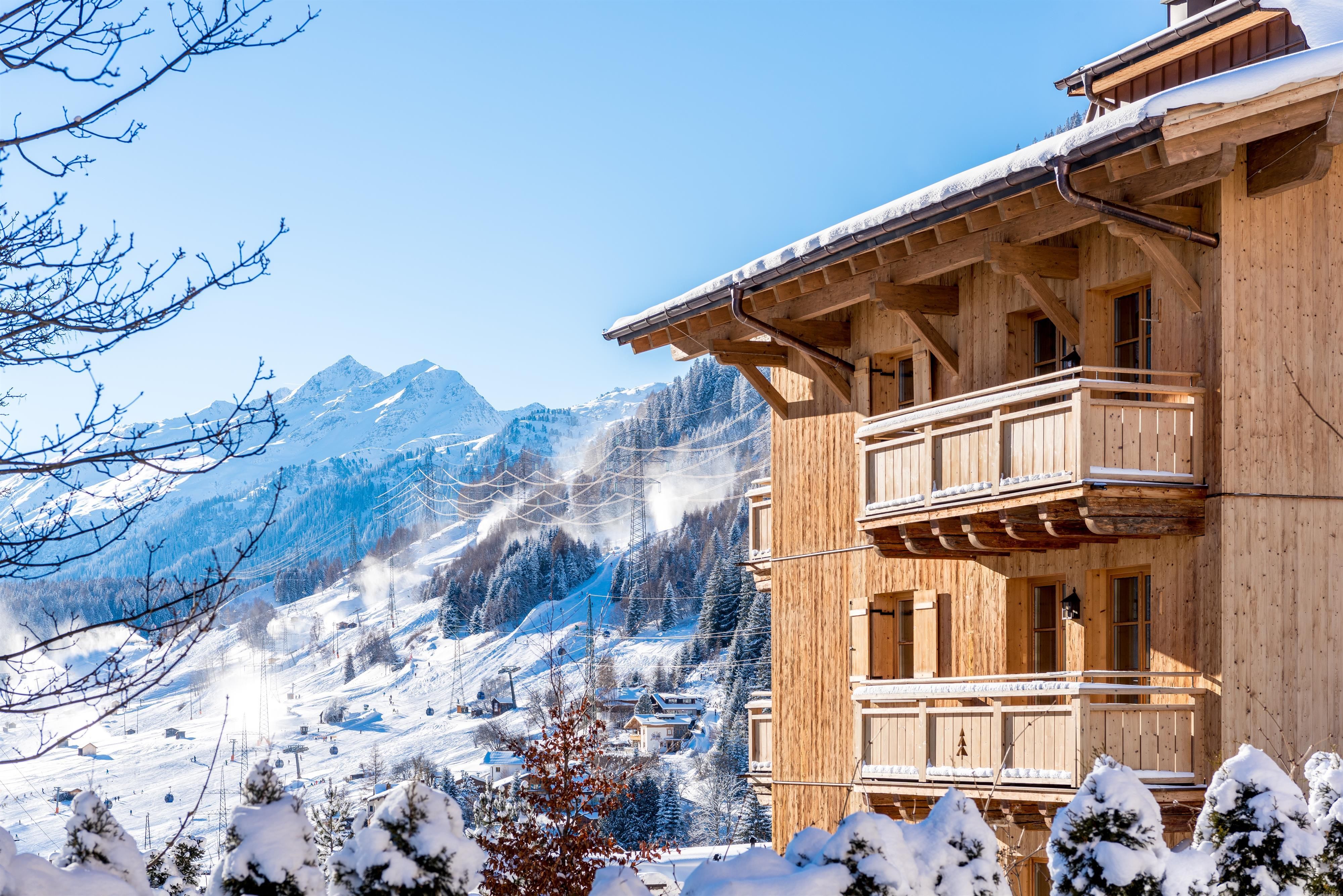 A cozy wooden building in the snow with balconies. In the background, there are snow-covered mountains and a clear blue sky.