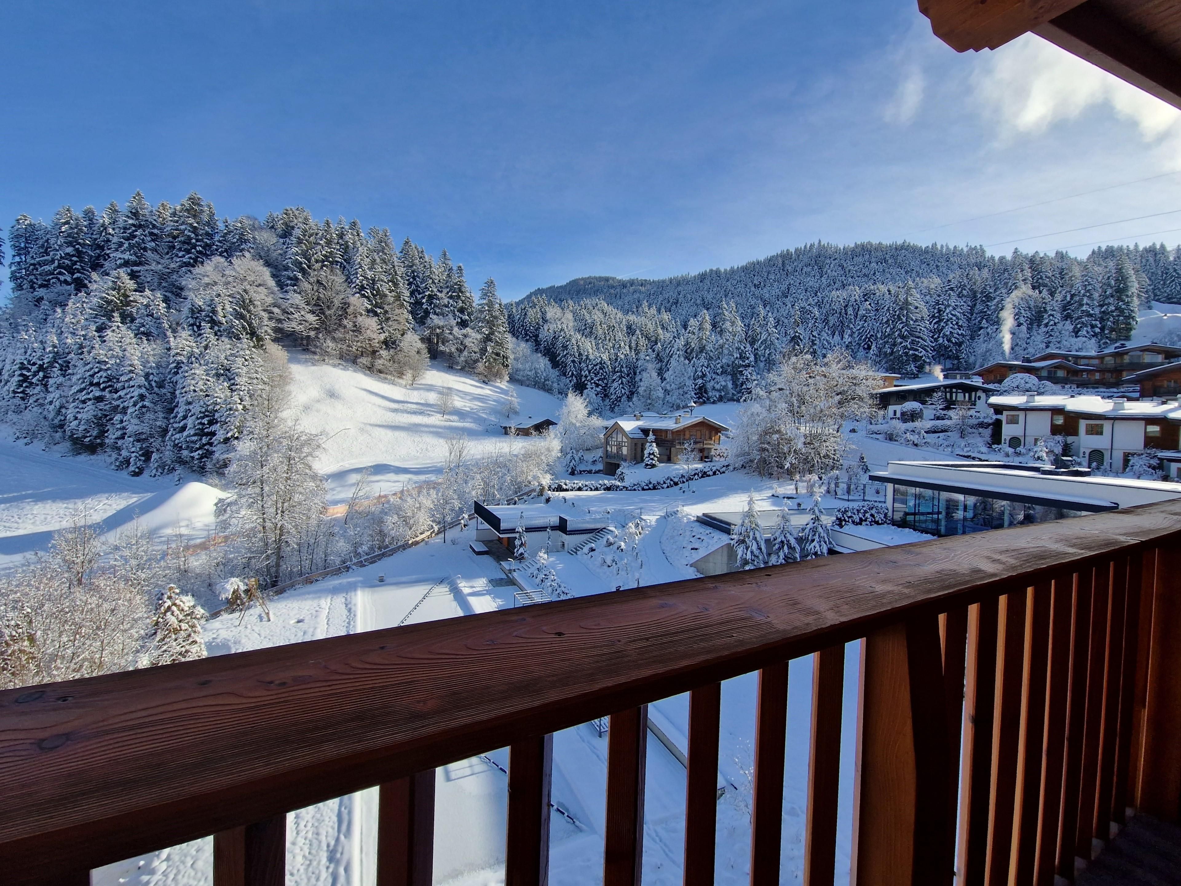 A snowy landscape with snow-covered trees and hills. In the foreground, a wooden balustrade can be seen.