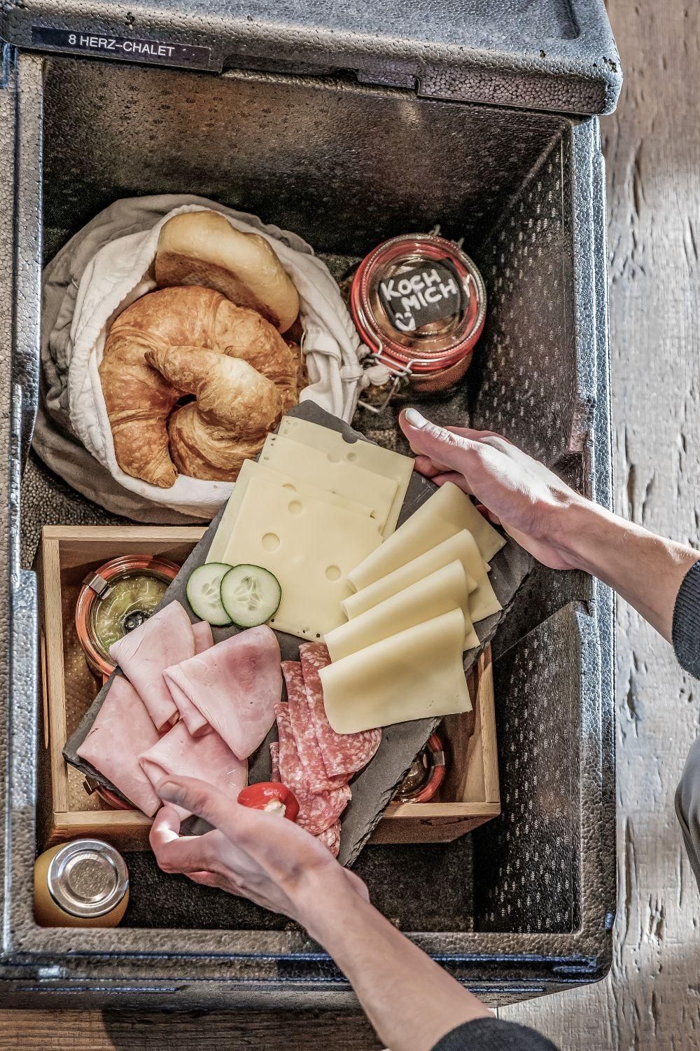 Breakfast box filled with fresh foods, including croissant, sausage, cheese, and vegetables. It also includes jars of jam and a drink.