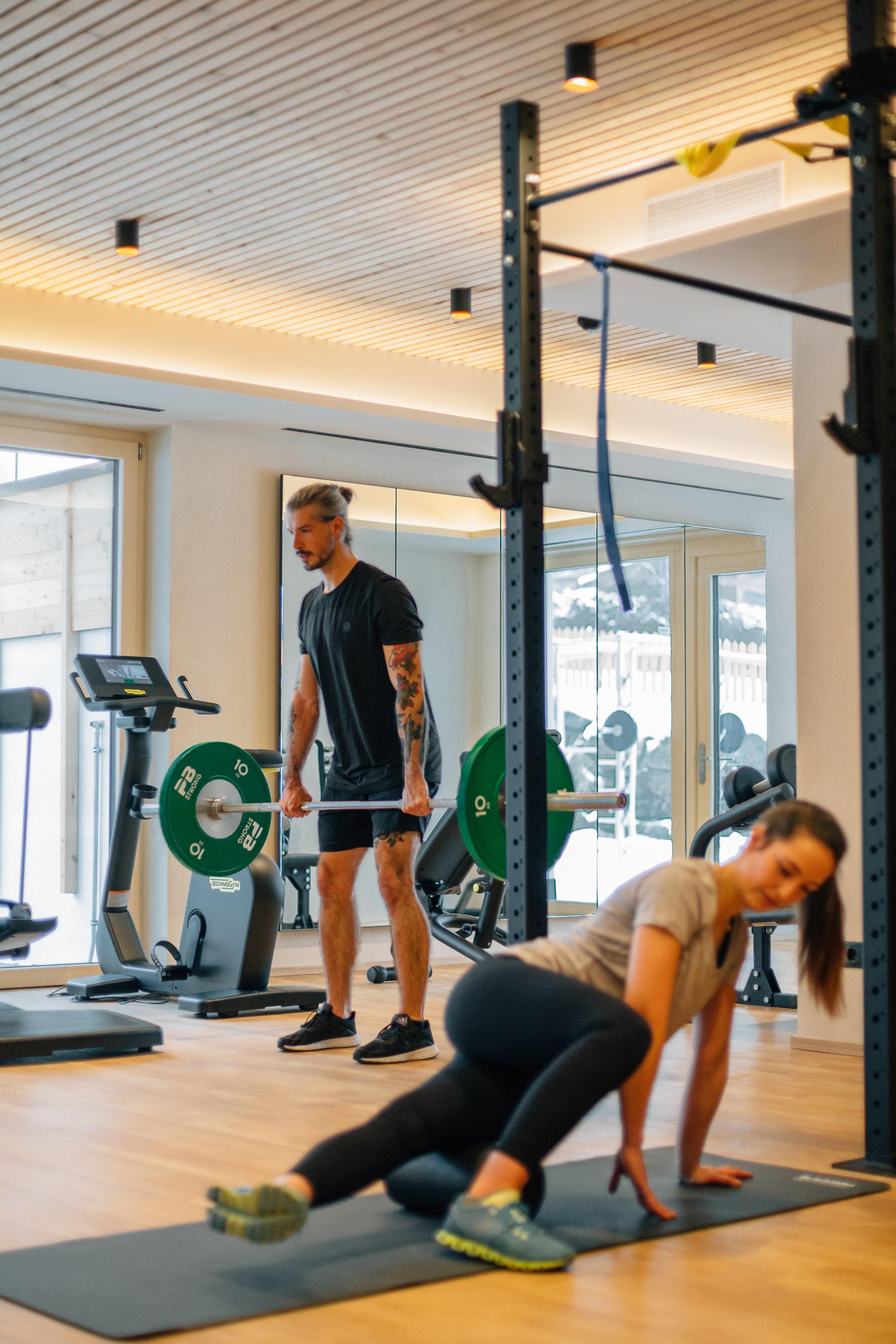 A modern gym environment with two people training. One person is doing an exercise on the floor while the other is watching in the background.