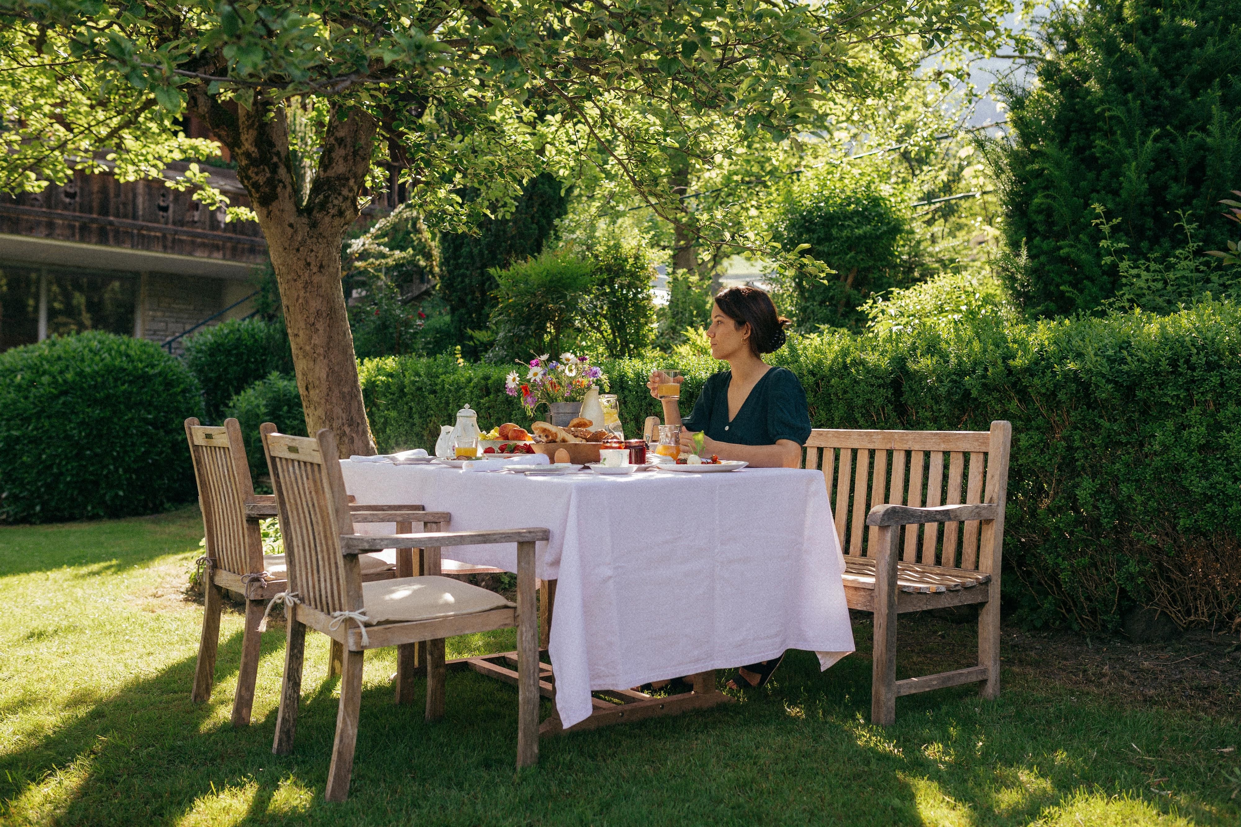 A beautiful garden with a set table and a woman eating relaxed. Surrounded by green bushes and trees, the scene radiates tranquility and harmony.