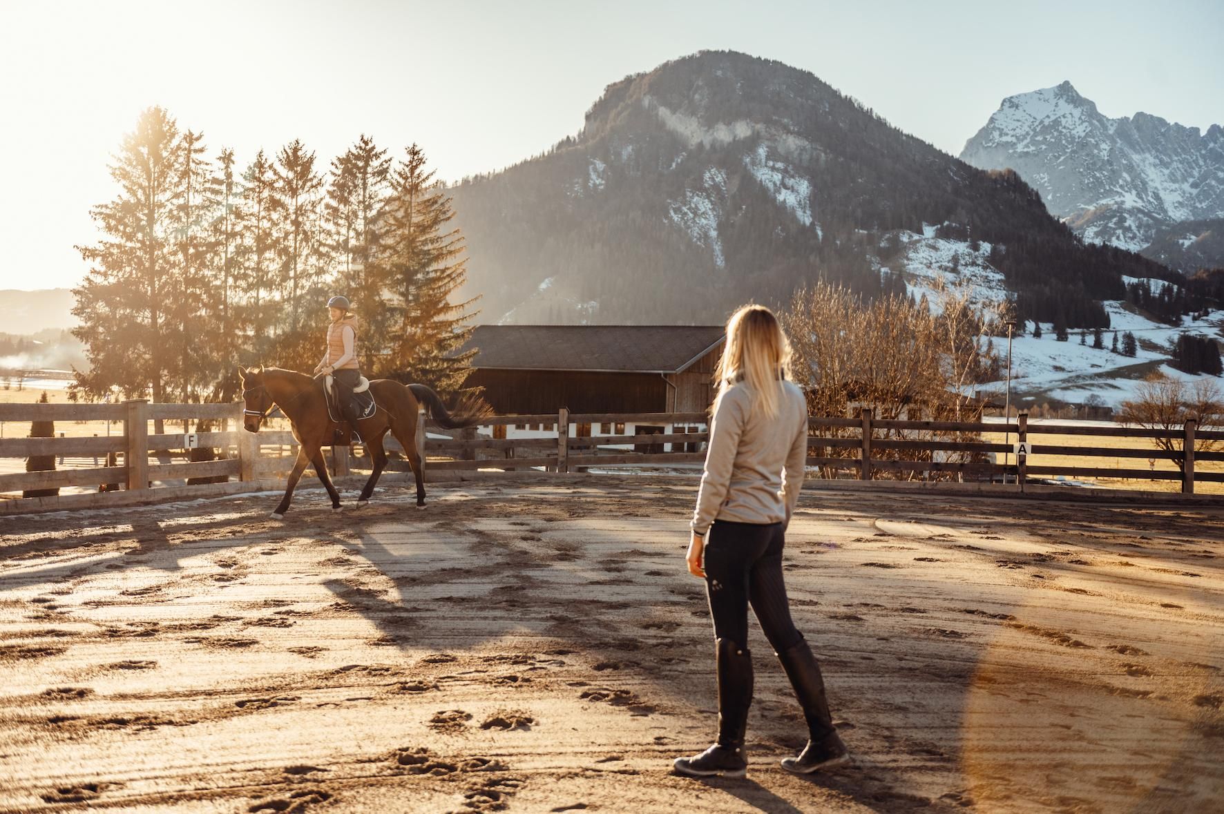 A person is riding a horse in a sandy arena. In the background, mountains and trees are visible as the sun sets.