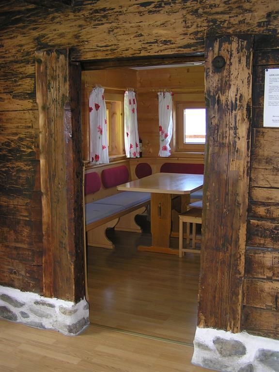 An inviting room with wooden paneling and a large table. Through the door frame, the simple, traditional furniture and windows with white curtains are visible.
