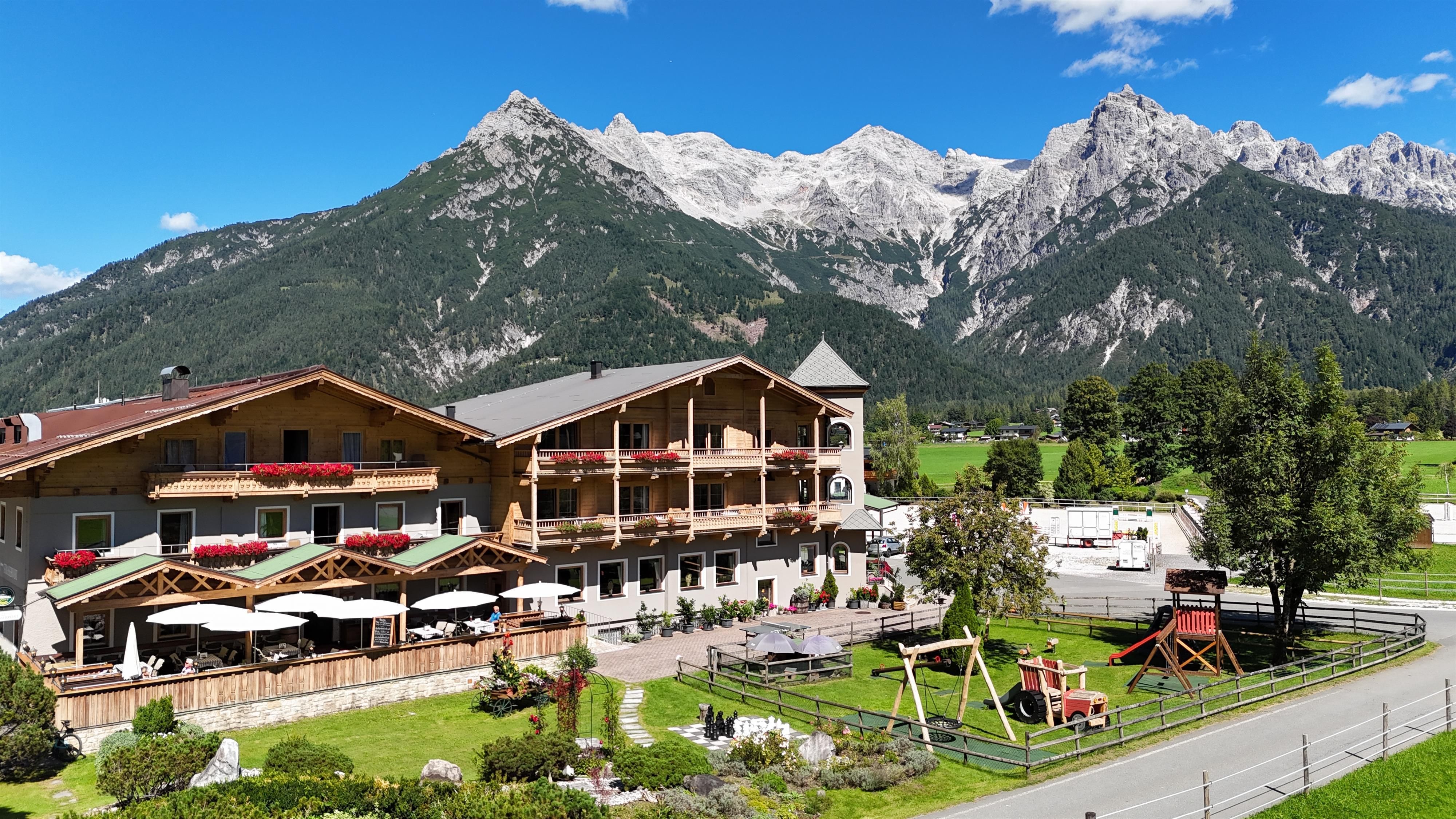 A picturesque hotel with a terrace and playground, surrounded by majestic mountains. The sky is clear and blue, ideal for a relaxing stay in nature.