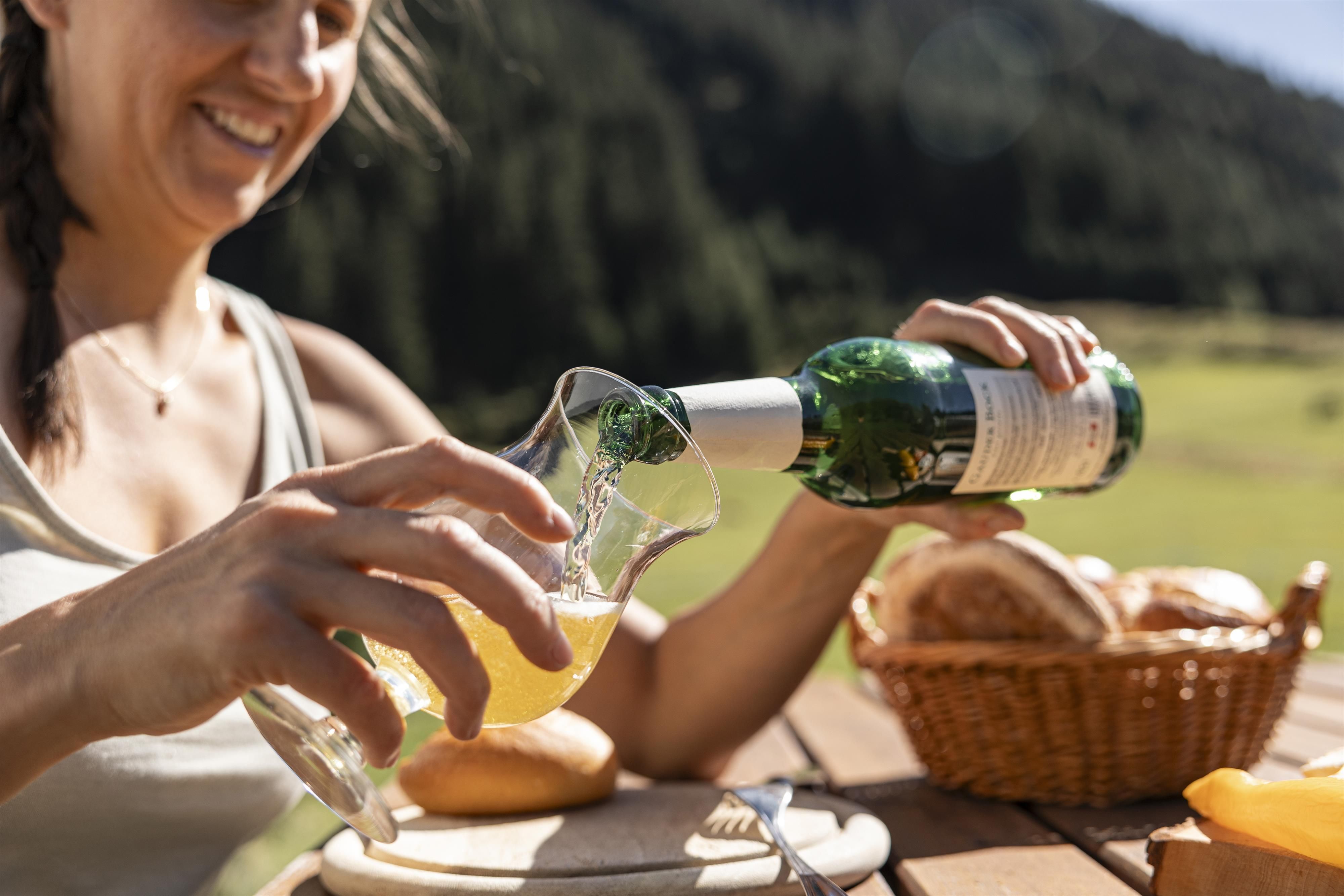 A woman is pouring a drink from a bottle into a glass. In the background, a green landscape and a basket with rolls can be seen.