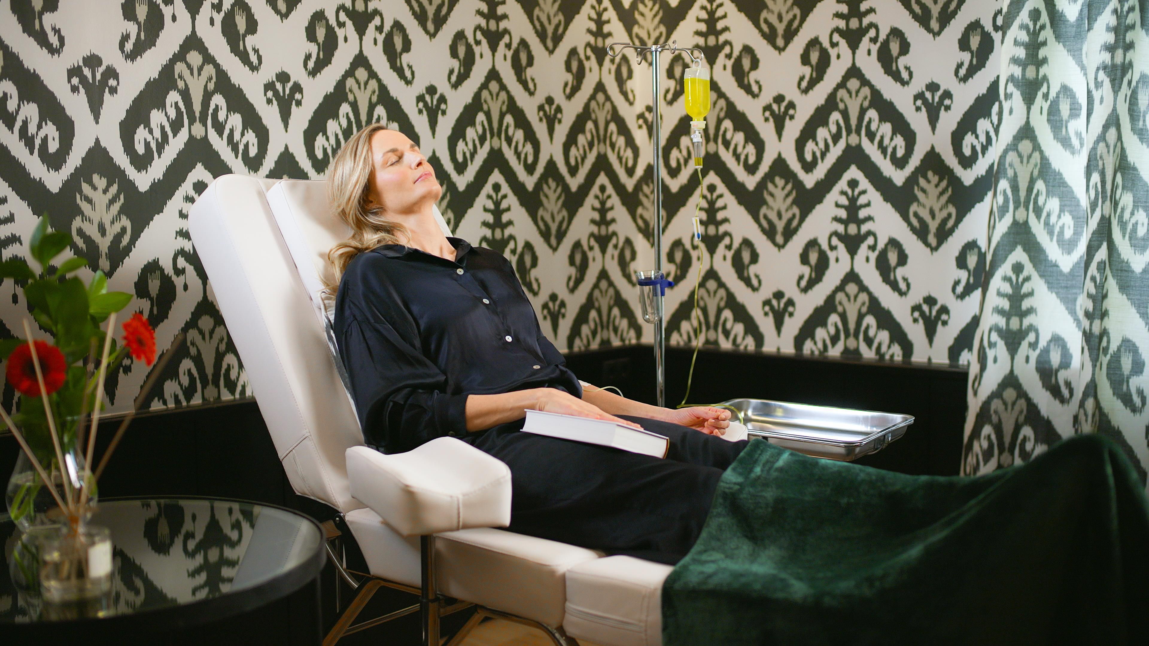 A relaxed woman sits in a comfortable chair and is receiving an infusion. The walls are decorated with an elegant pattern.
