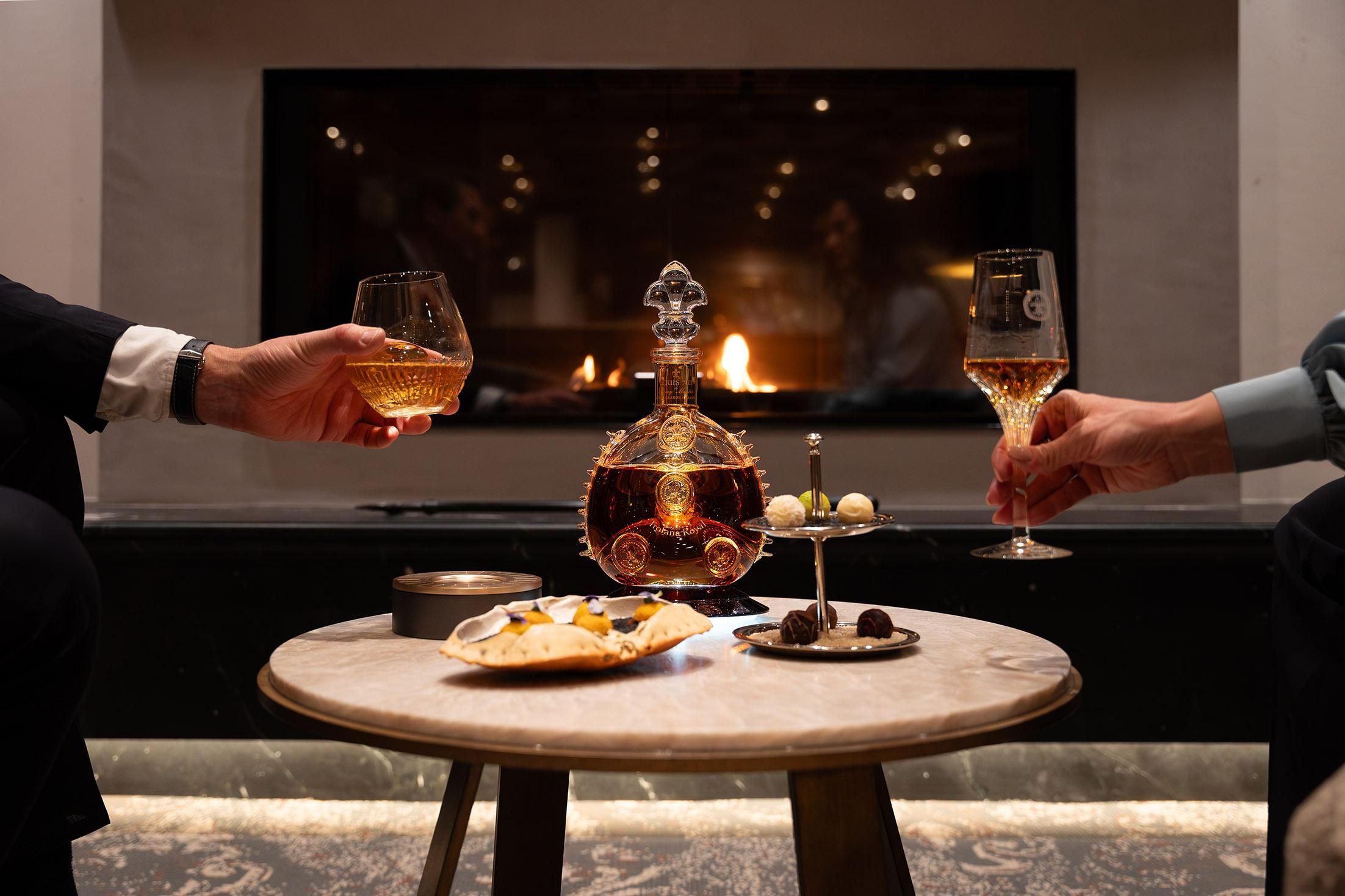 Two people clink glasses while sitting at the table. In the background, a fireplace is burning, and snacks and an elegant carafe are on the table.