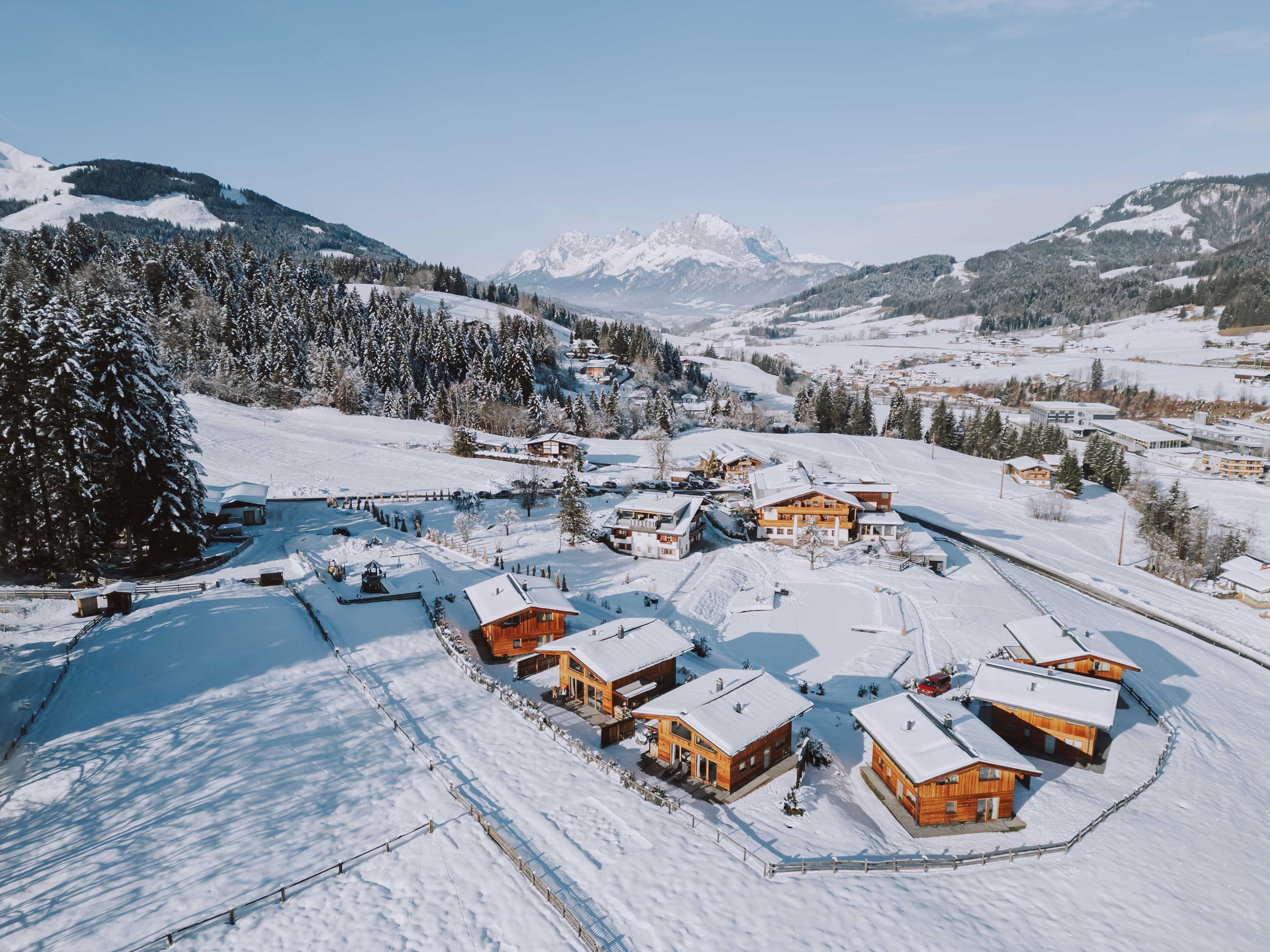 A picturesque winter landscape with snow-covered cabins. In the background, mountains and forests are visible.
