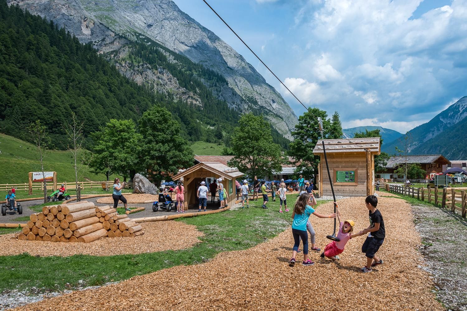 A playground in nature with swinging children and wooden elements. In the background, there are mountains and a group of people visible.