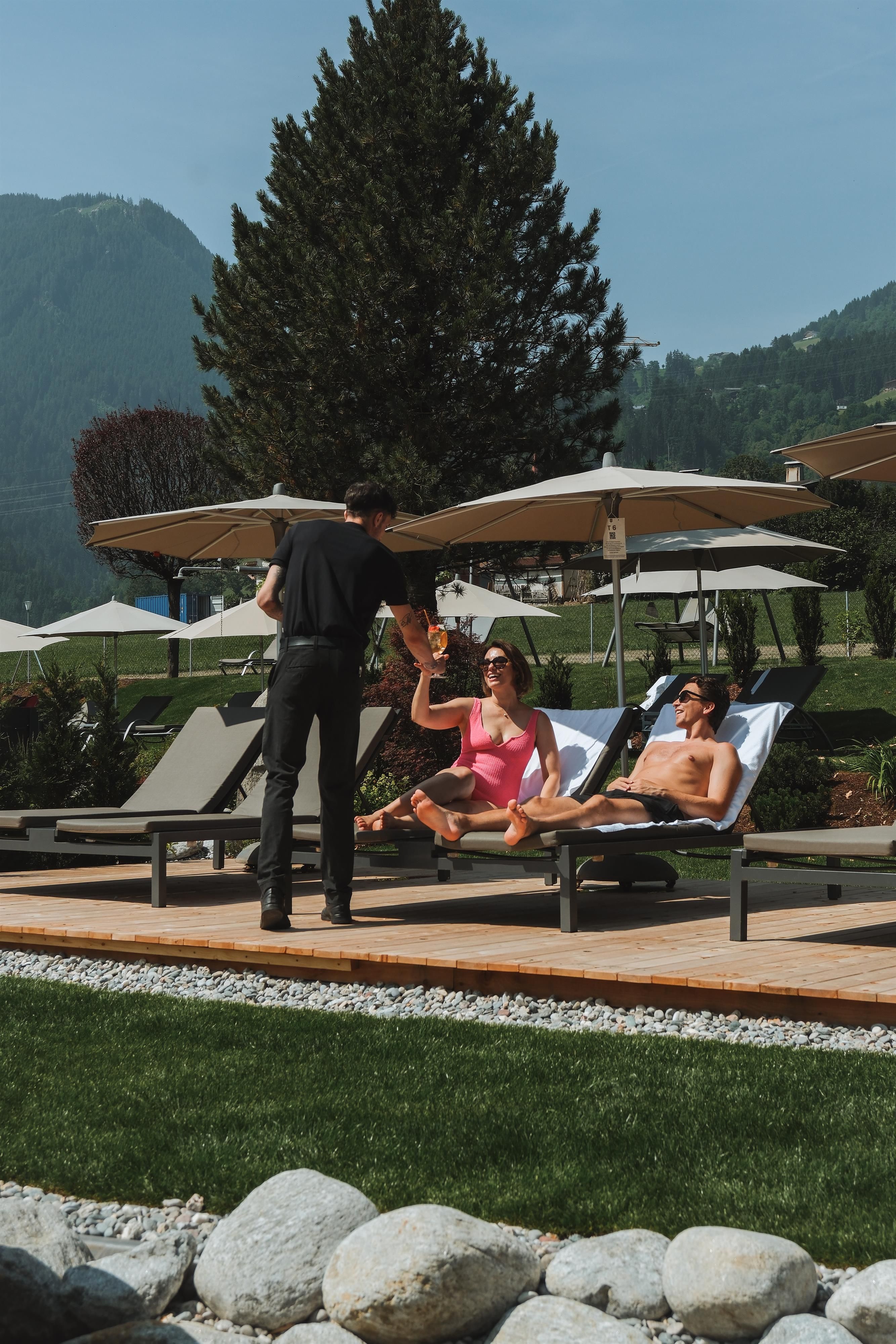 A sunny pool area with loungers and umbrellas. Two people are relaxing and getting drinks from a waiter.