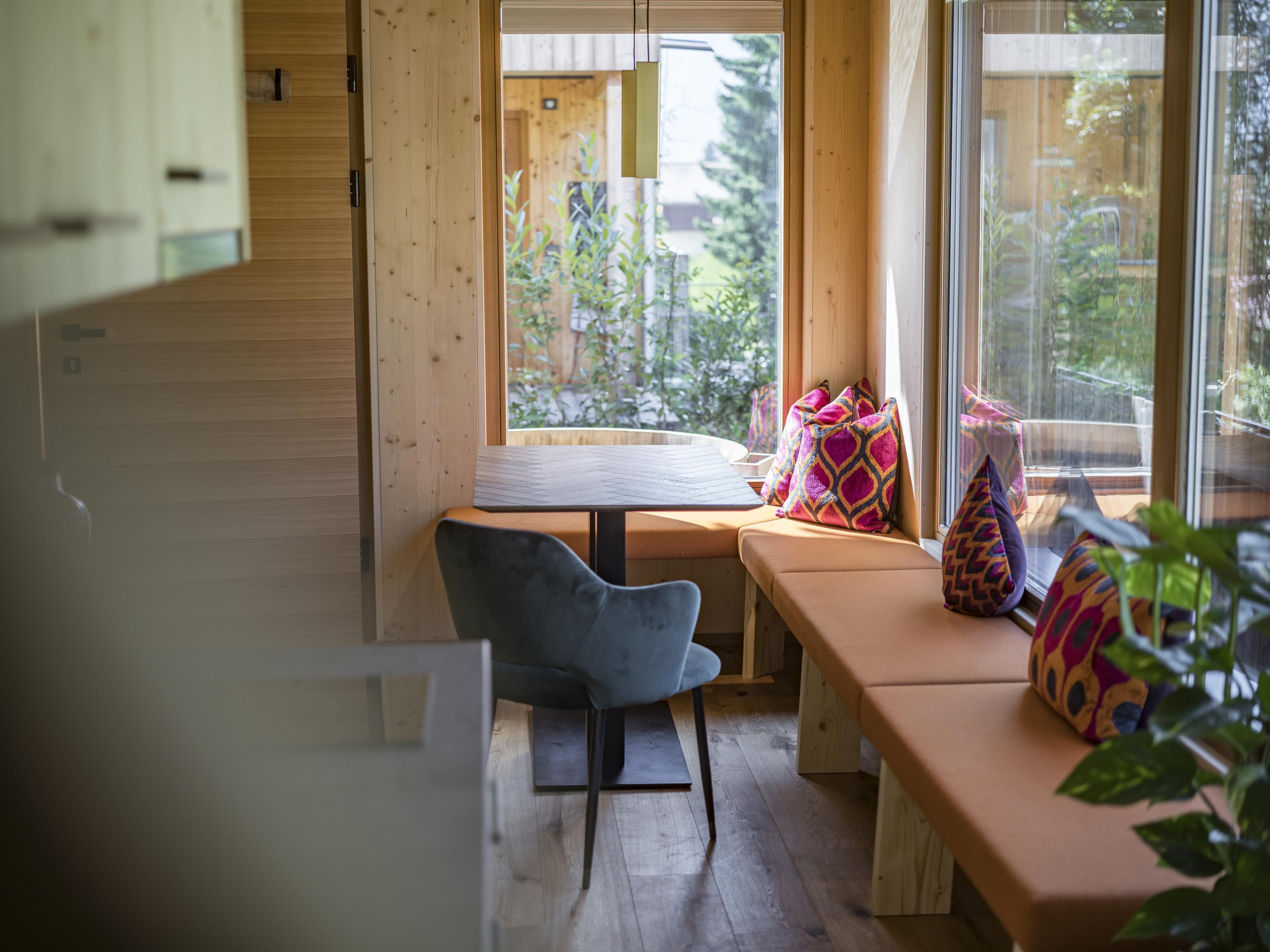 A bright cozy seating area with a table and colorful cushions. Large windows offer a view of the outdoor area.