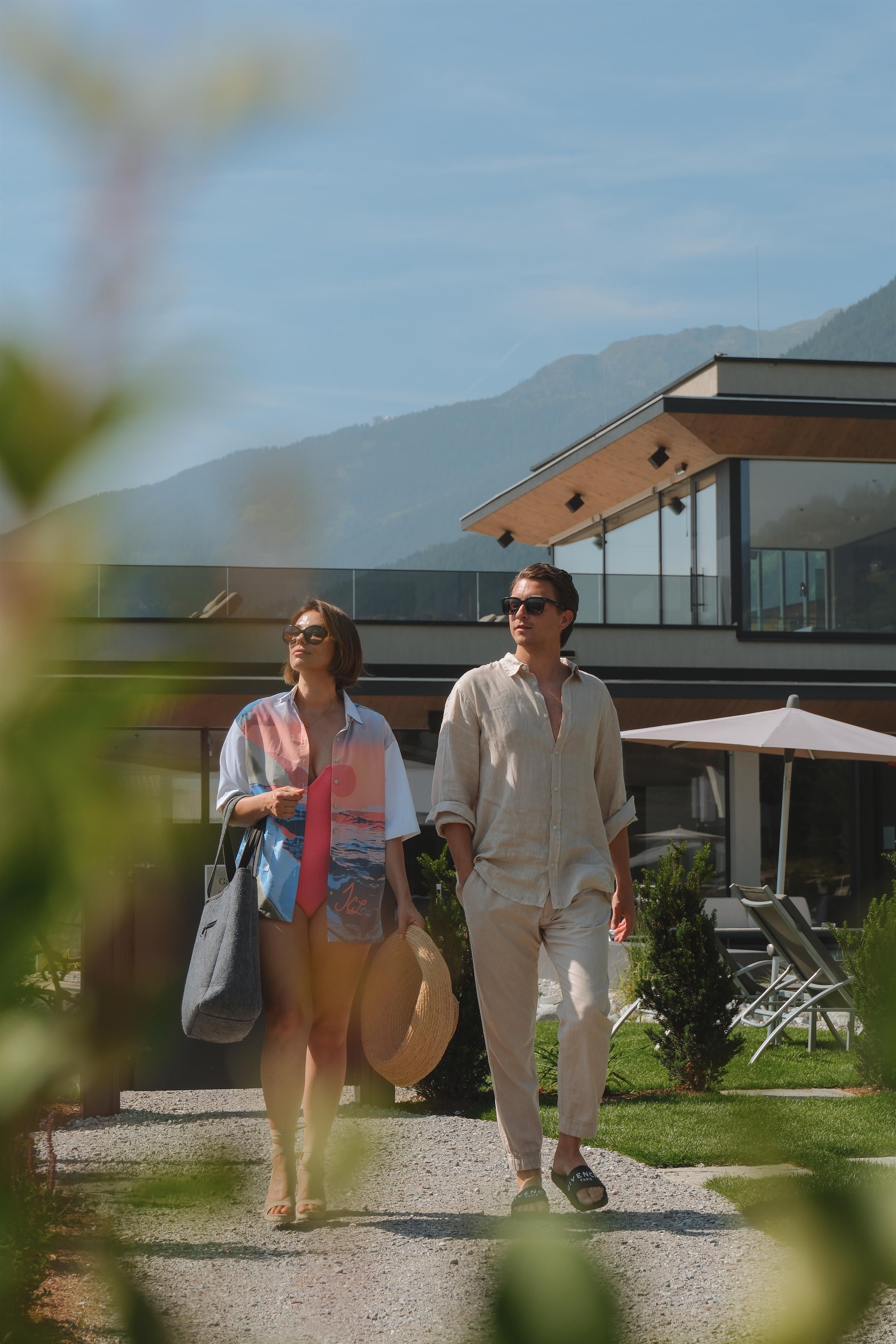 A couple is walking in a modern garden. In the background, there is an elegant building with mountains and a blue sky.