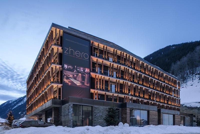 A modern hotel in the mountains, surrounded by snow. The facade is designed with wood and large windows, and there is a striking advertisement board.