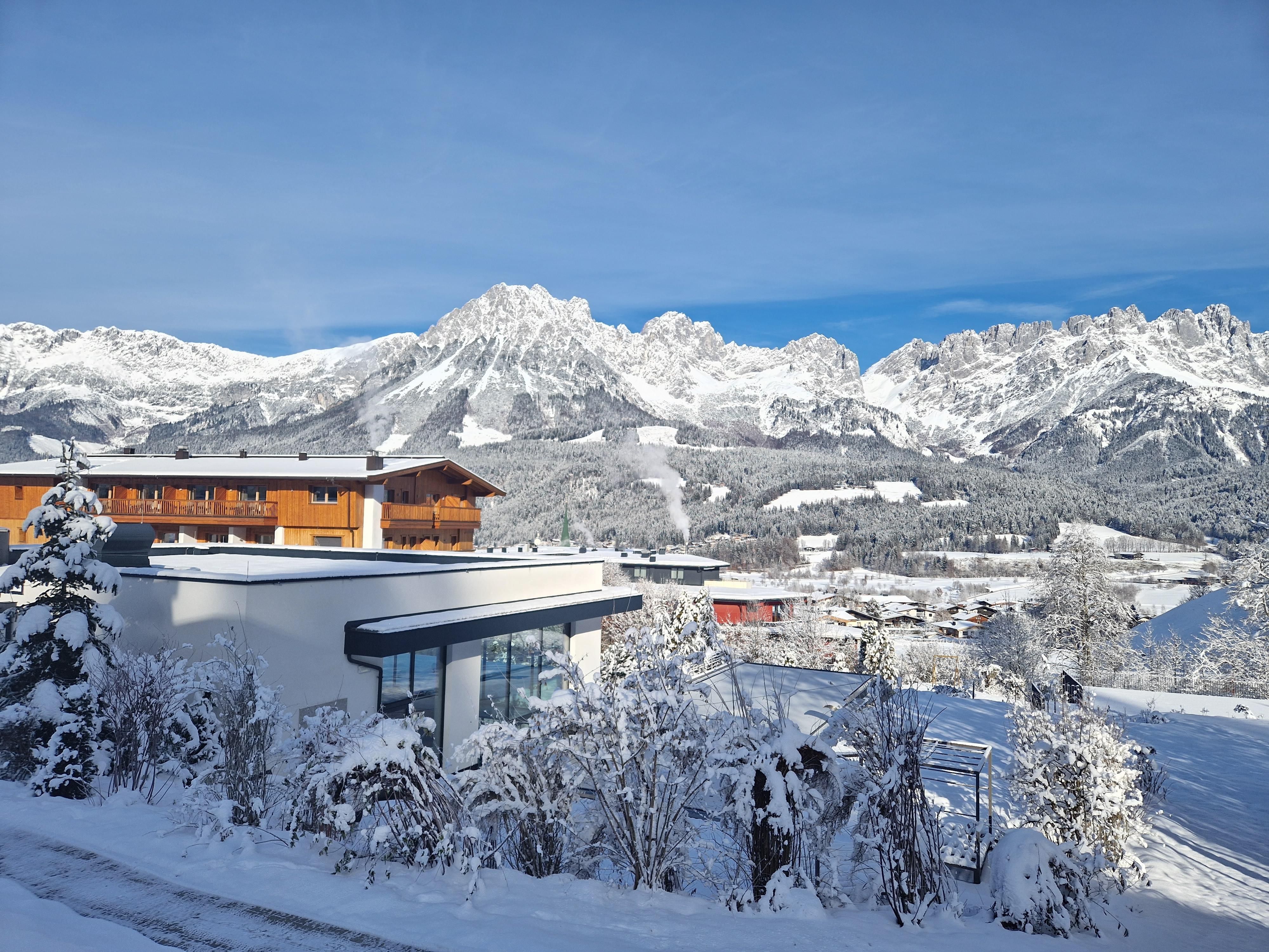 A snowy landscape with majestic mountains in the background. In the foreground, modern buildings and snow-covered trees stand.