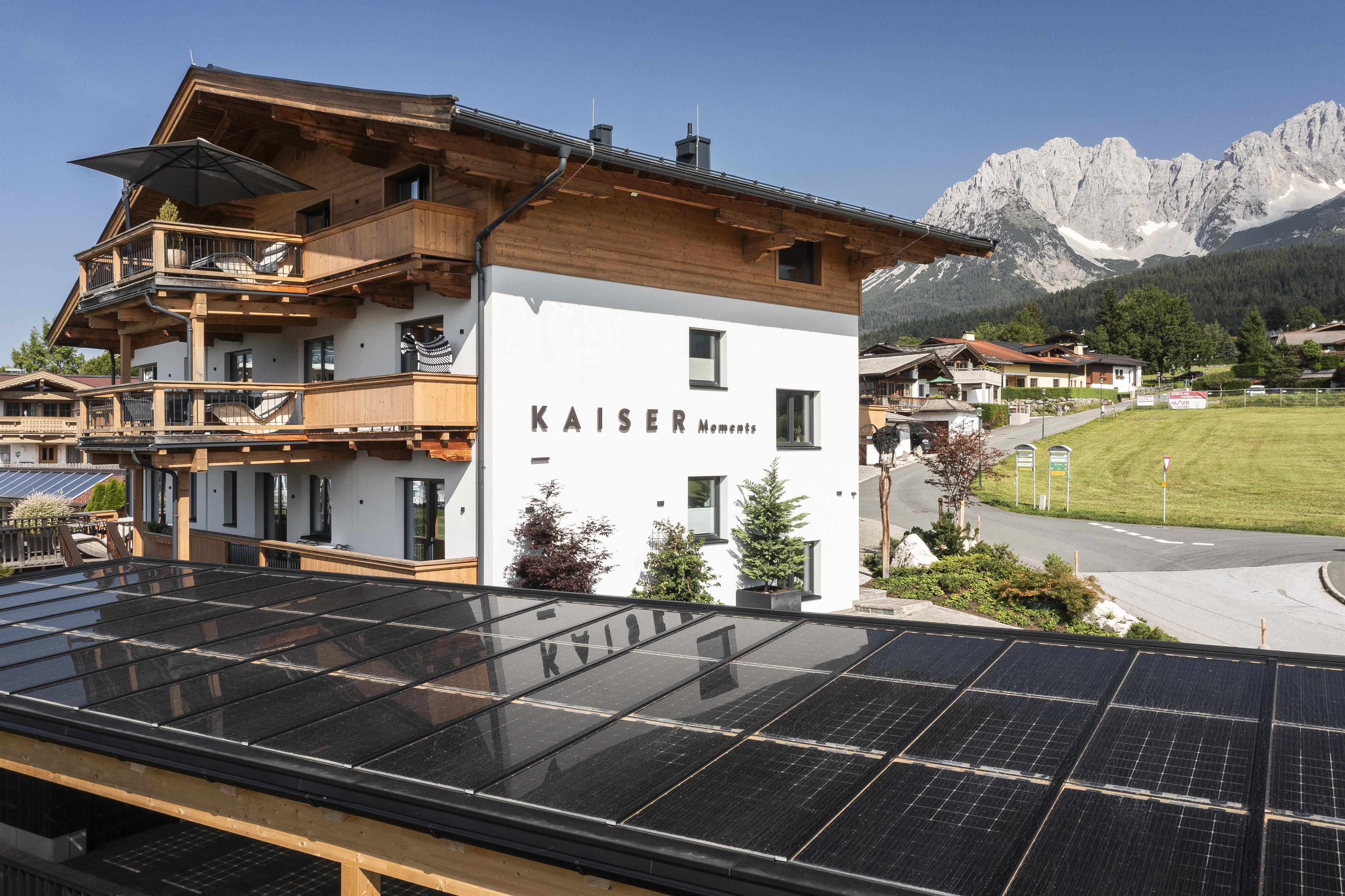 A modern building named "Kaiser" with balconies, surrounded by a beautiful mountain landscape. Solar panels on the roof complement the sustainable architecture.