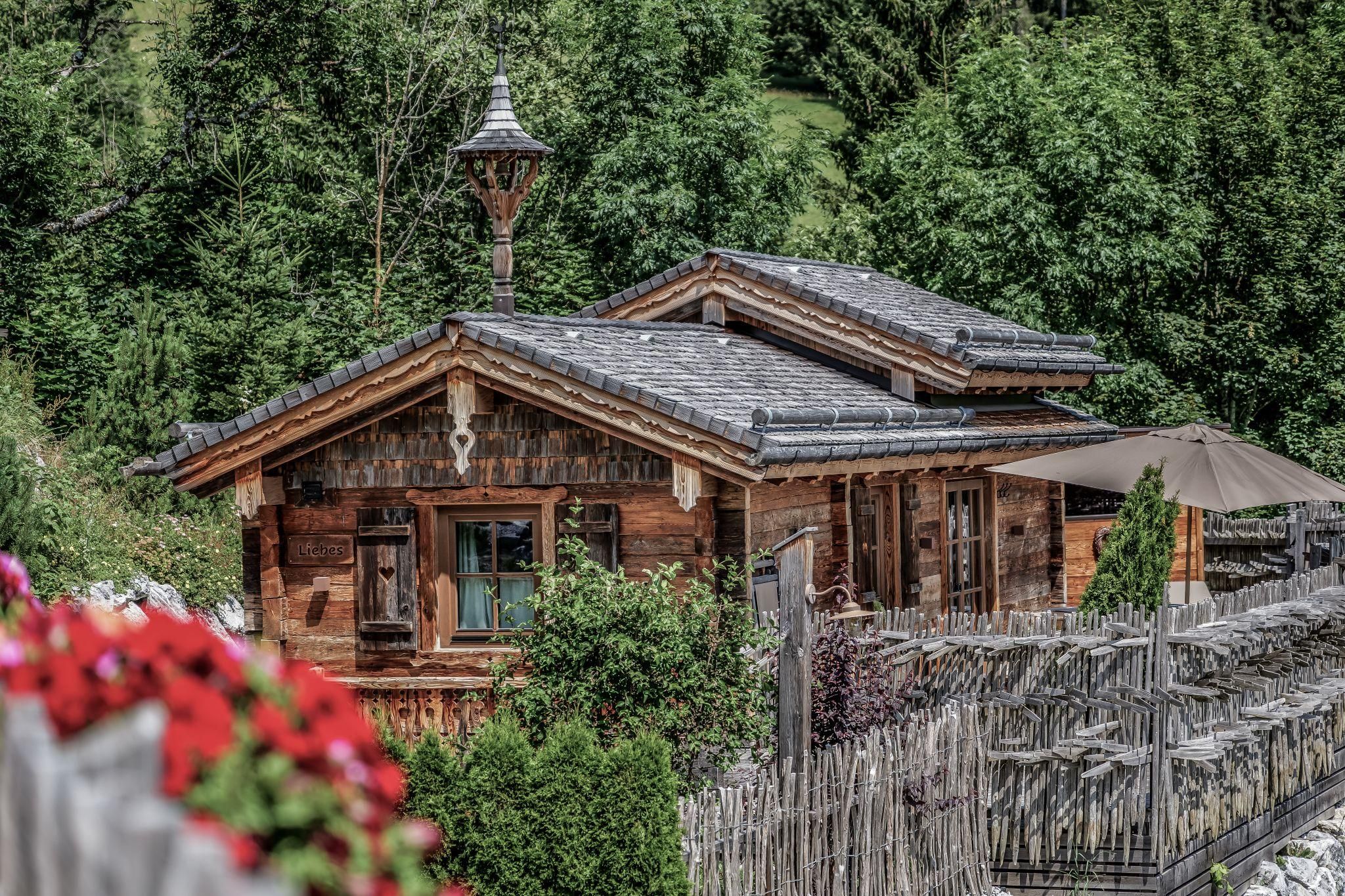 An idyllic chalet surrounded by green trees. Flowers and a parasol give the scene an inviting charm.