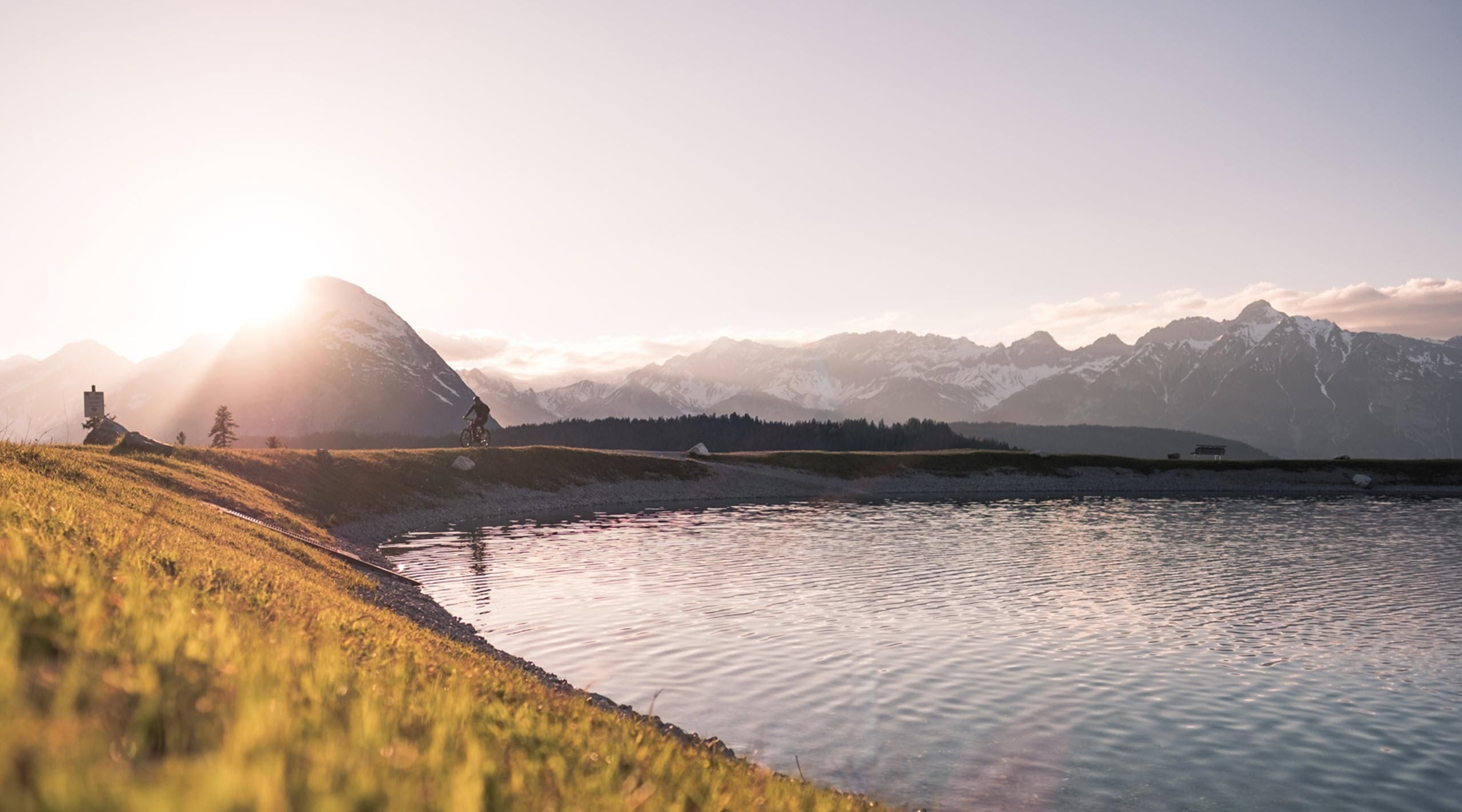 A peaceful landscape with a lake and mountains in the background. The sun rises behind the mountains, casting warm light over the scene.