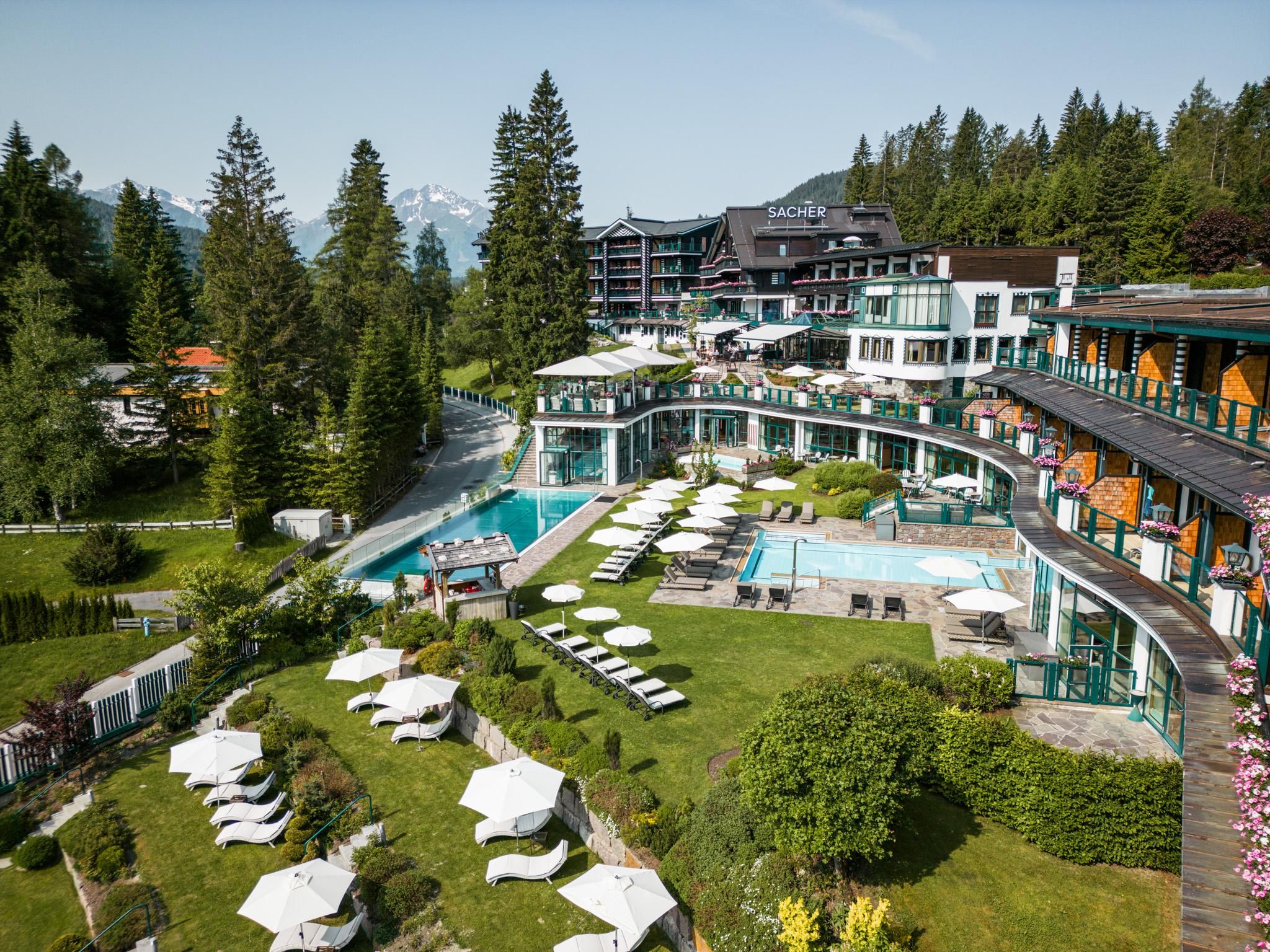 A luxurious hotel with a pool area and expansive green spaces. Surrounding are tall trees, and the mountains in the background are visible.