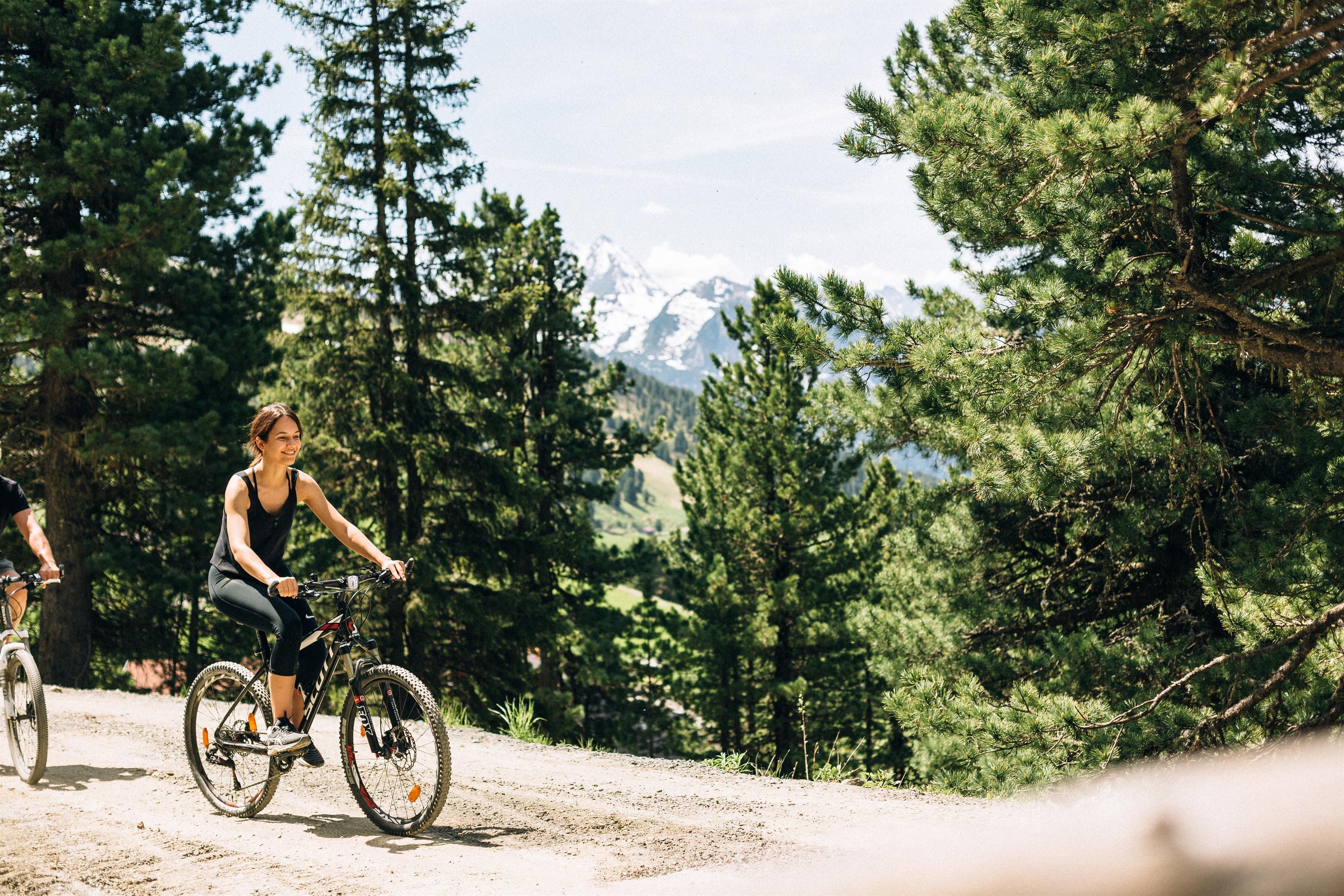 A woman is riding a bicycle on a path through a forest. In the background, mountains and a clear sky can be seen.