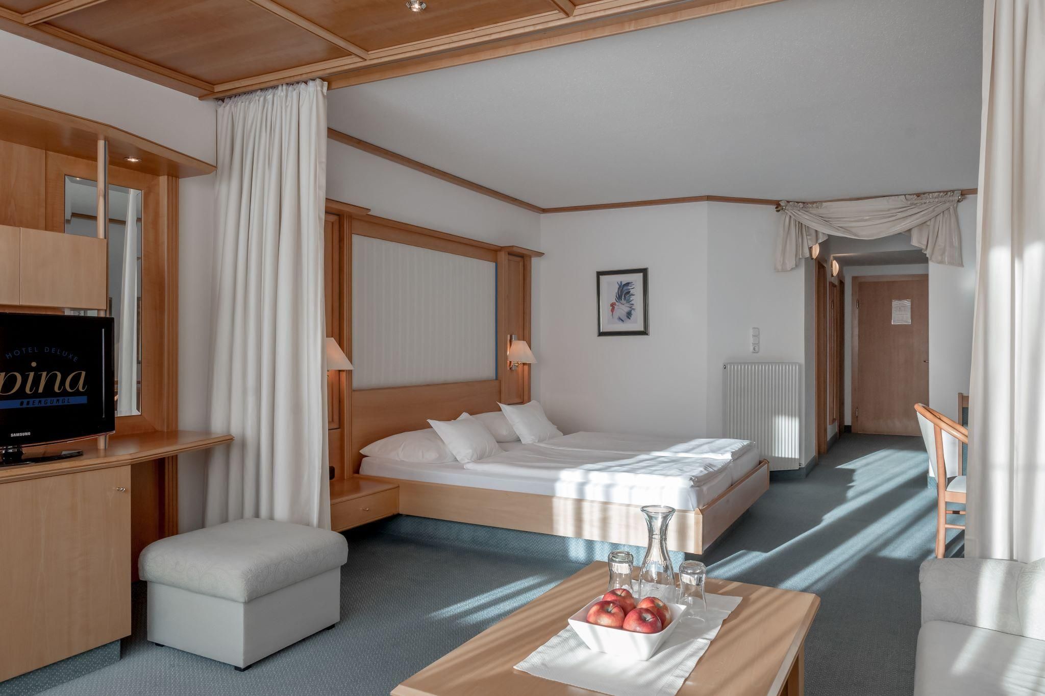 A modern hotel room with a double bed and wooden furniture. There is a small table with fruit and a TV in the corner.