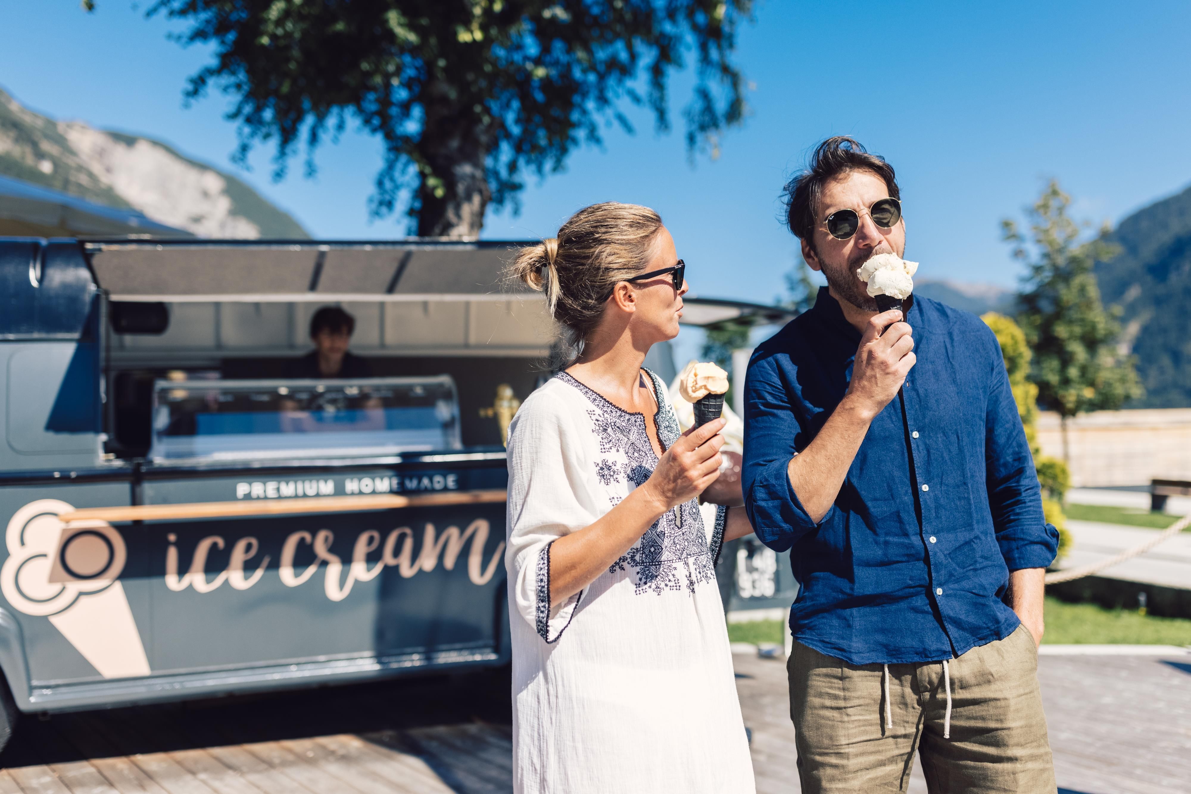 A couple enjoys ice cream in front of an ice cream truck in nature. In the background, mountains and trees are visible.