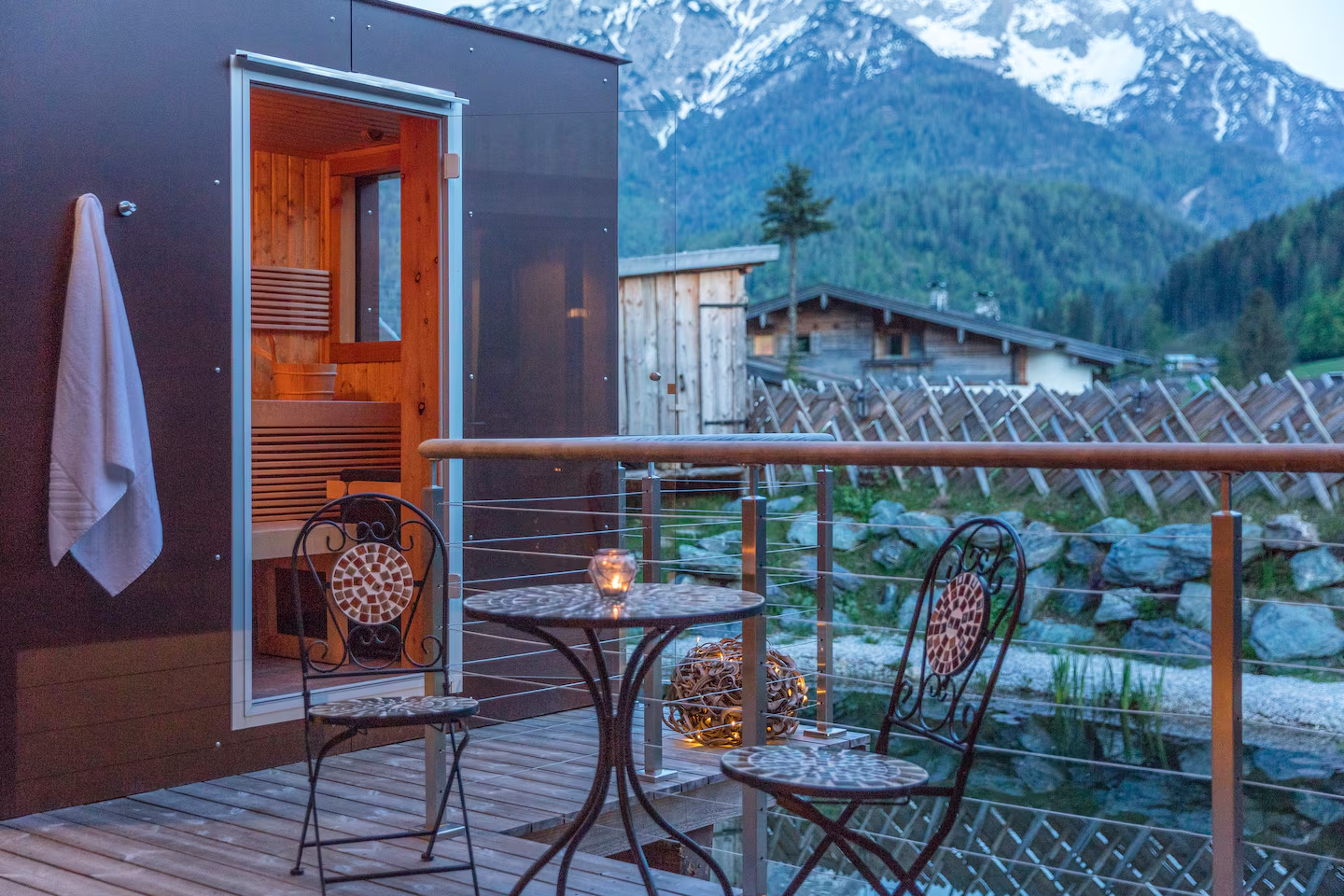 A cozy terrace with two chairs and a table. In the background, there are mountains and a sauna.