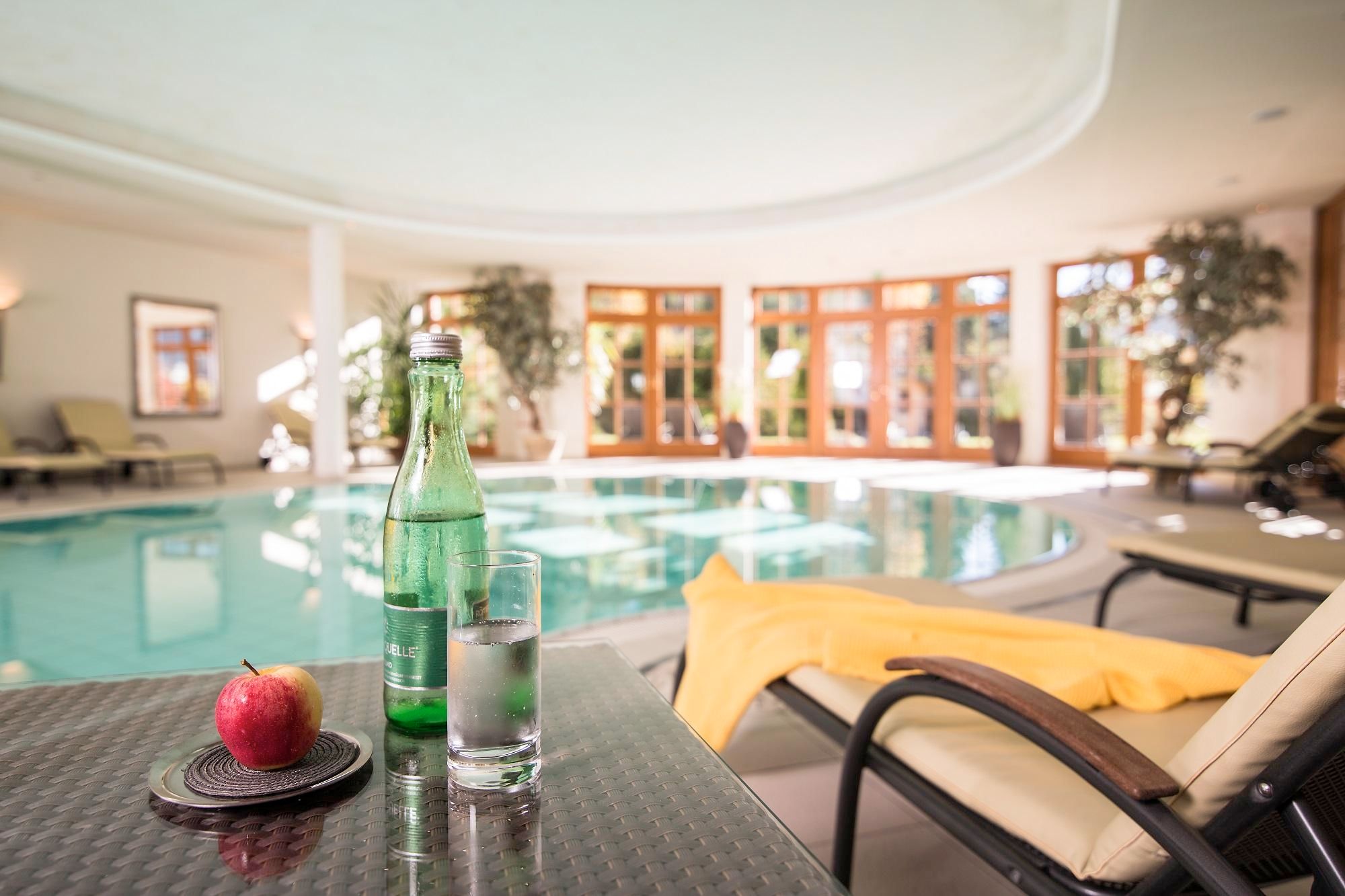 A relaxing indoor pool with spacious windows and natural light. On the table is a bottle of water, a glass, and a red apple.