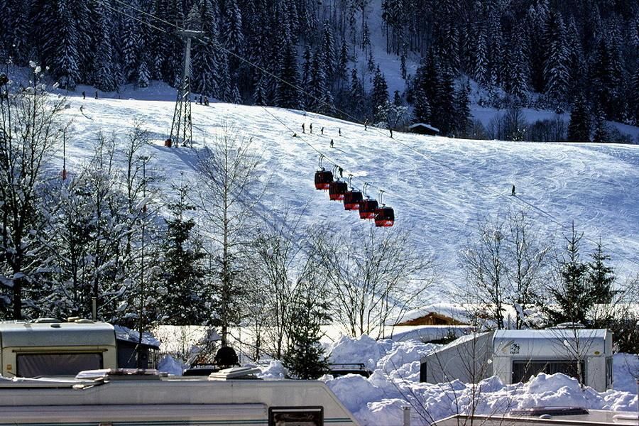 A snow-covered slope with a ski lift in the background. In the foreground, there are motorhomes and trees visible.