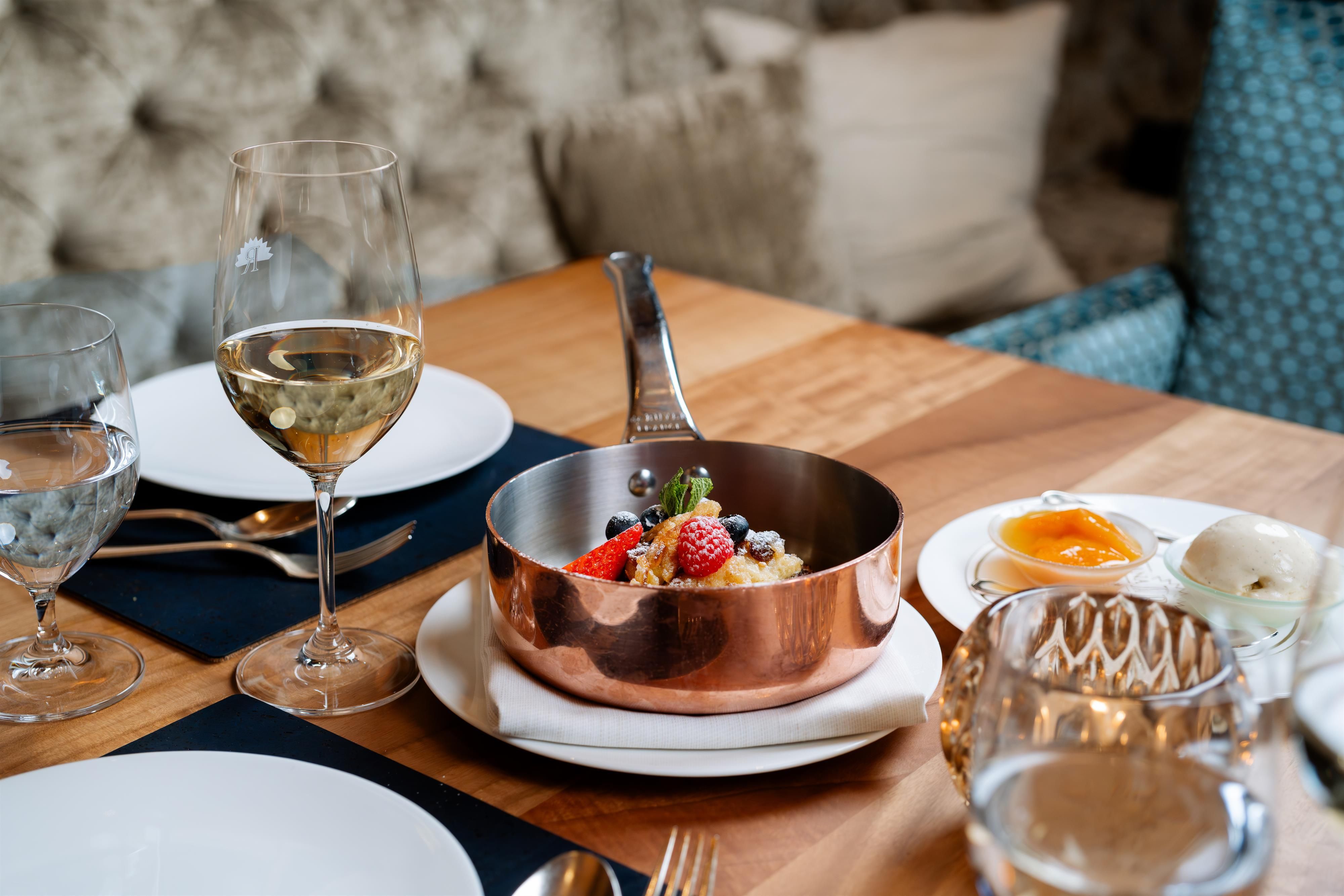 A stylishly set table with a copper container full of fresh berries and a serving of dessert. There are glasses of water and wine, as well as elegant cutlery.