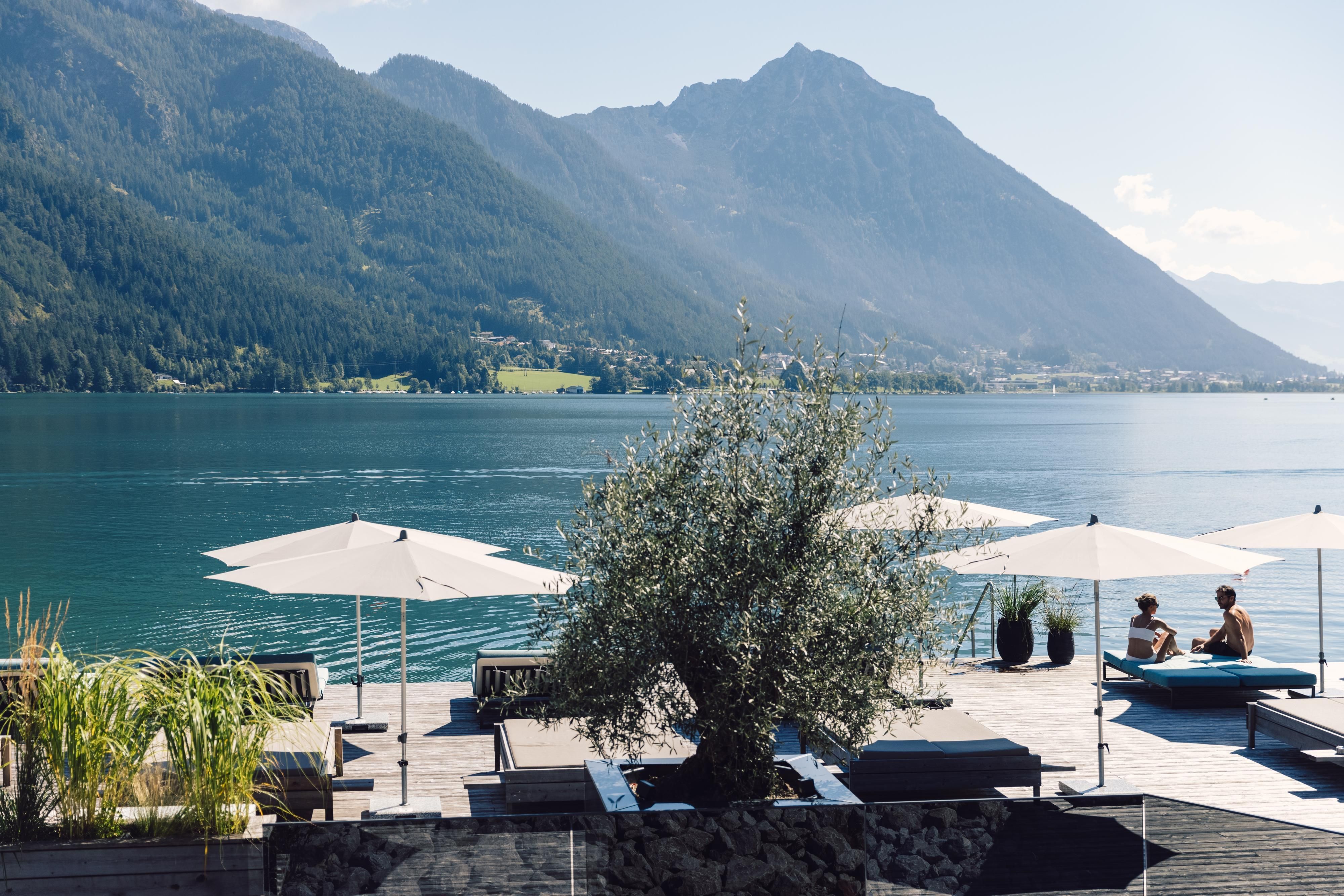 A beautiful lake with a view of mountains and a clear sky. Two people relax under beach umbrellas by the water.