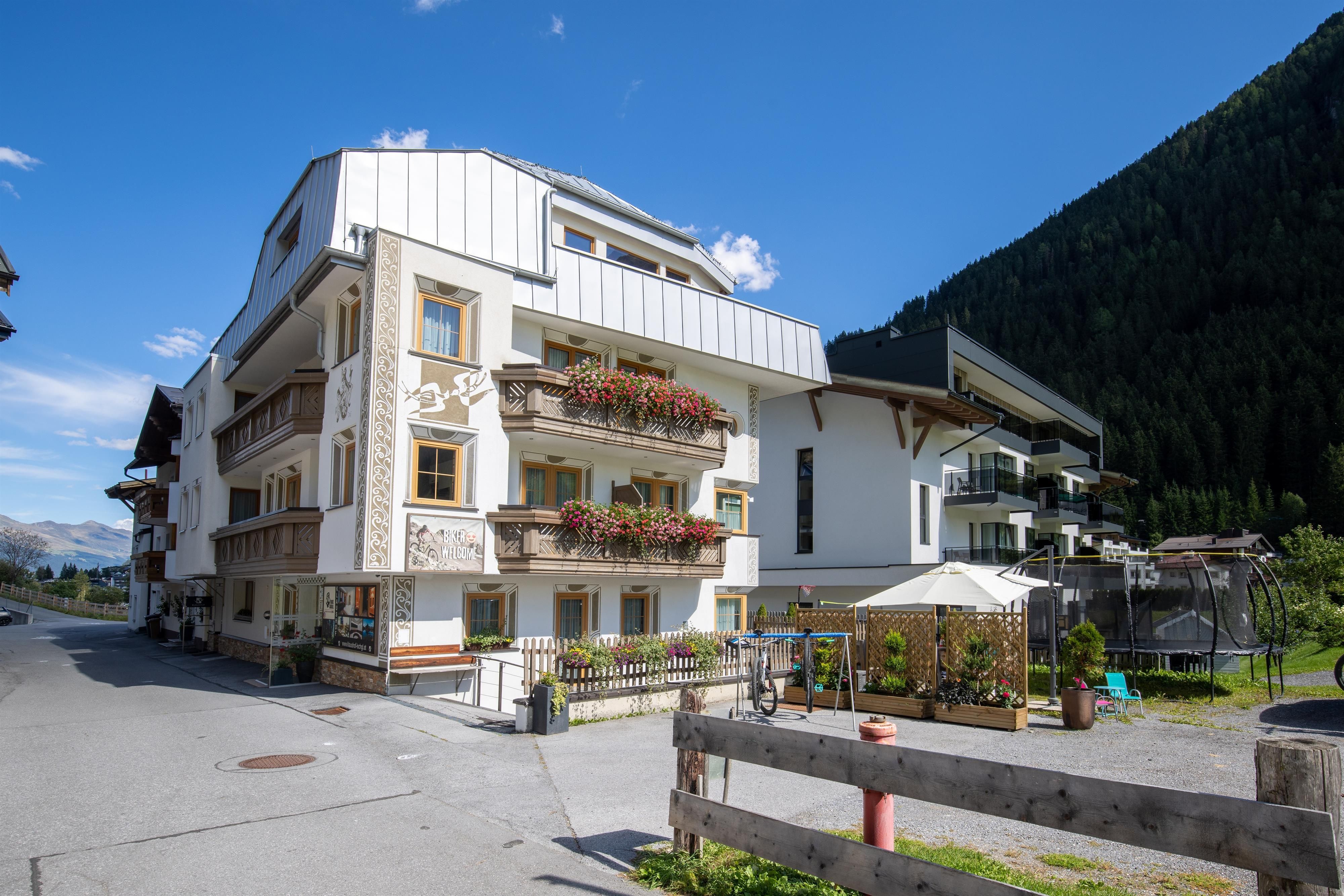 A modern building with balconies and flower arrangements. In the background, green mountains and a blue sky can be seen.