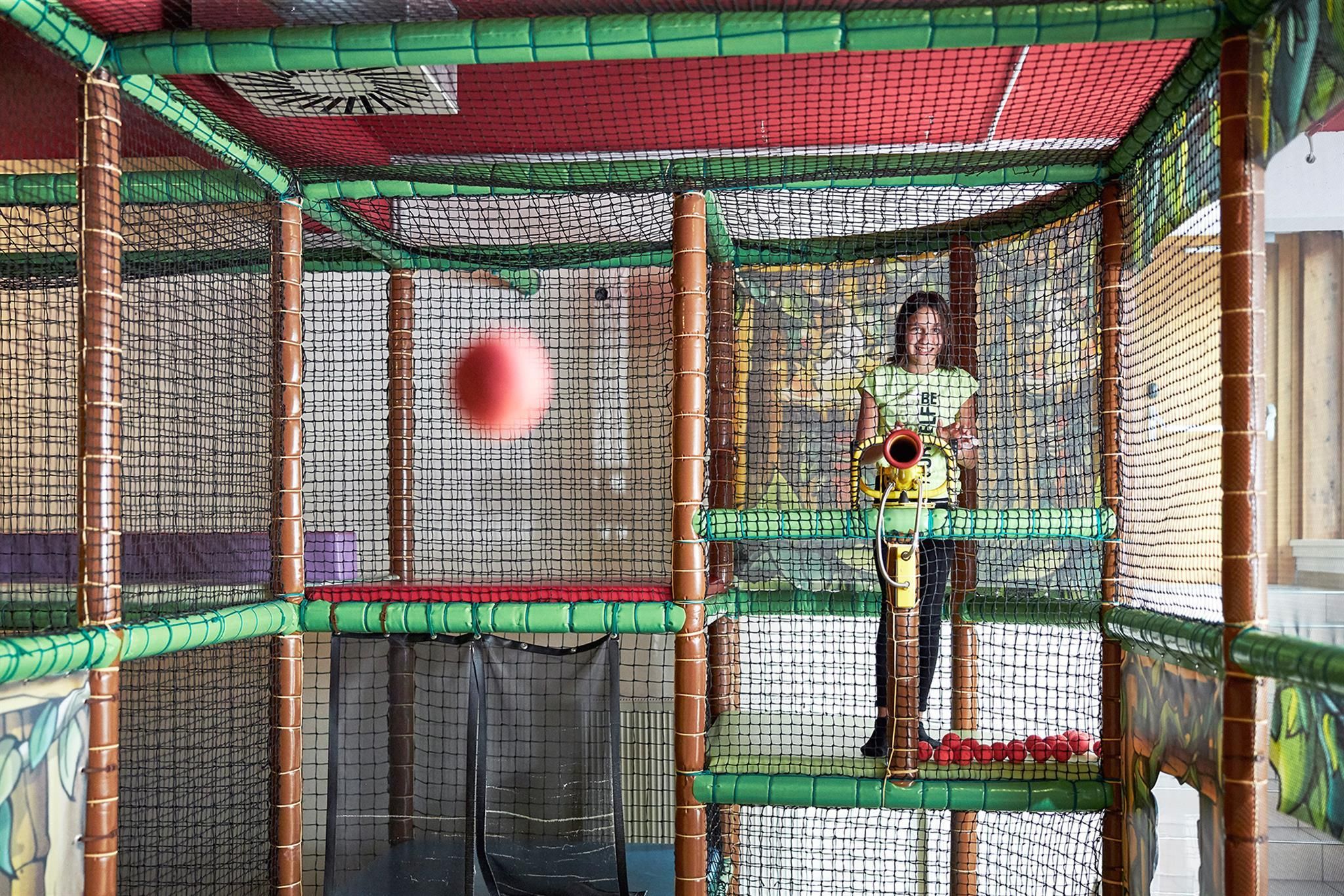 A play area with net structures and a child shooting a red ball with a toy. The environment is colorful and inviting.