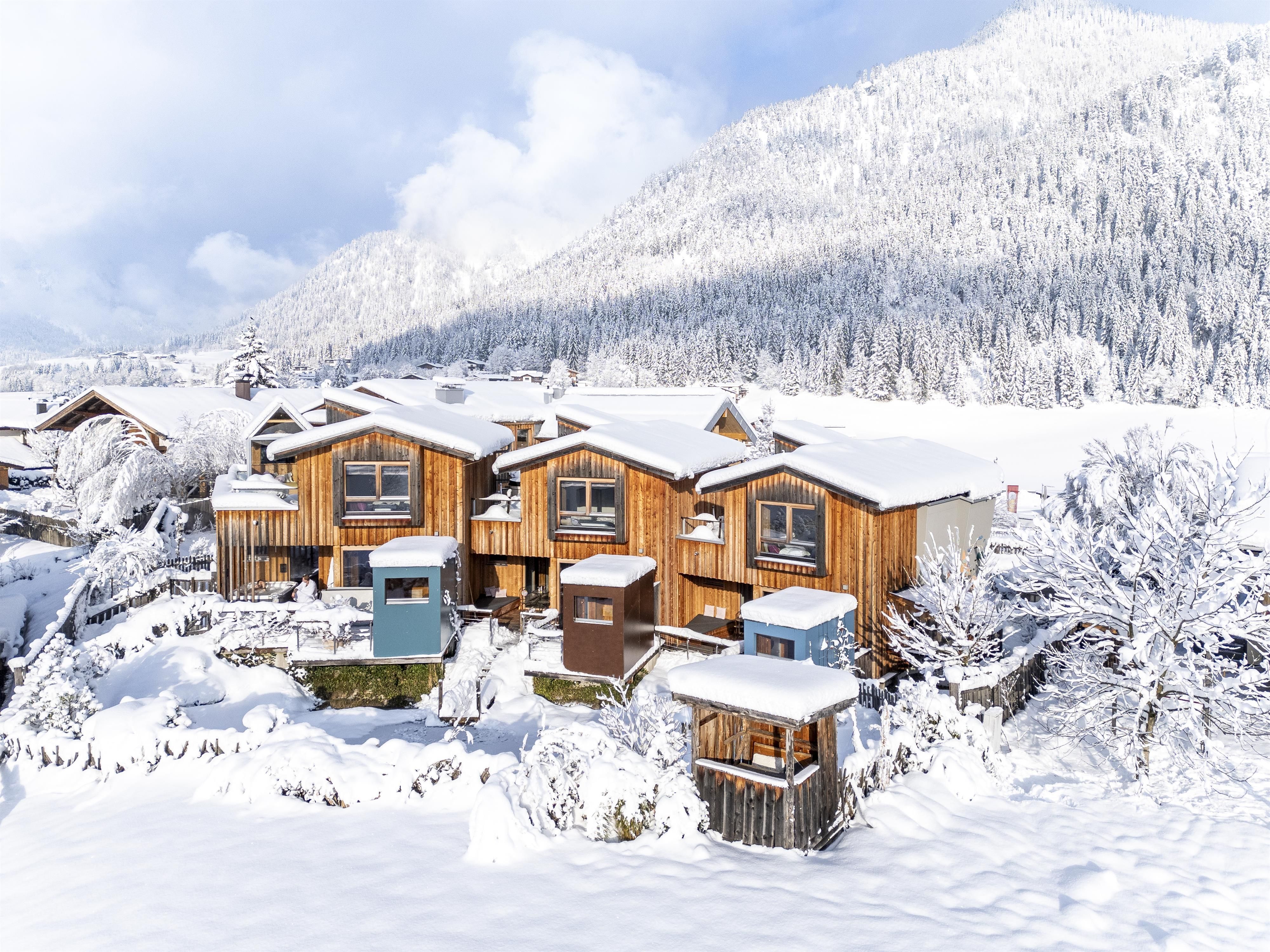 Snow-covered cabins in a winter landscape. In the background, high mountains and a clear blue sky can be seen.