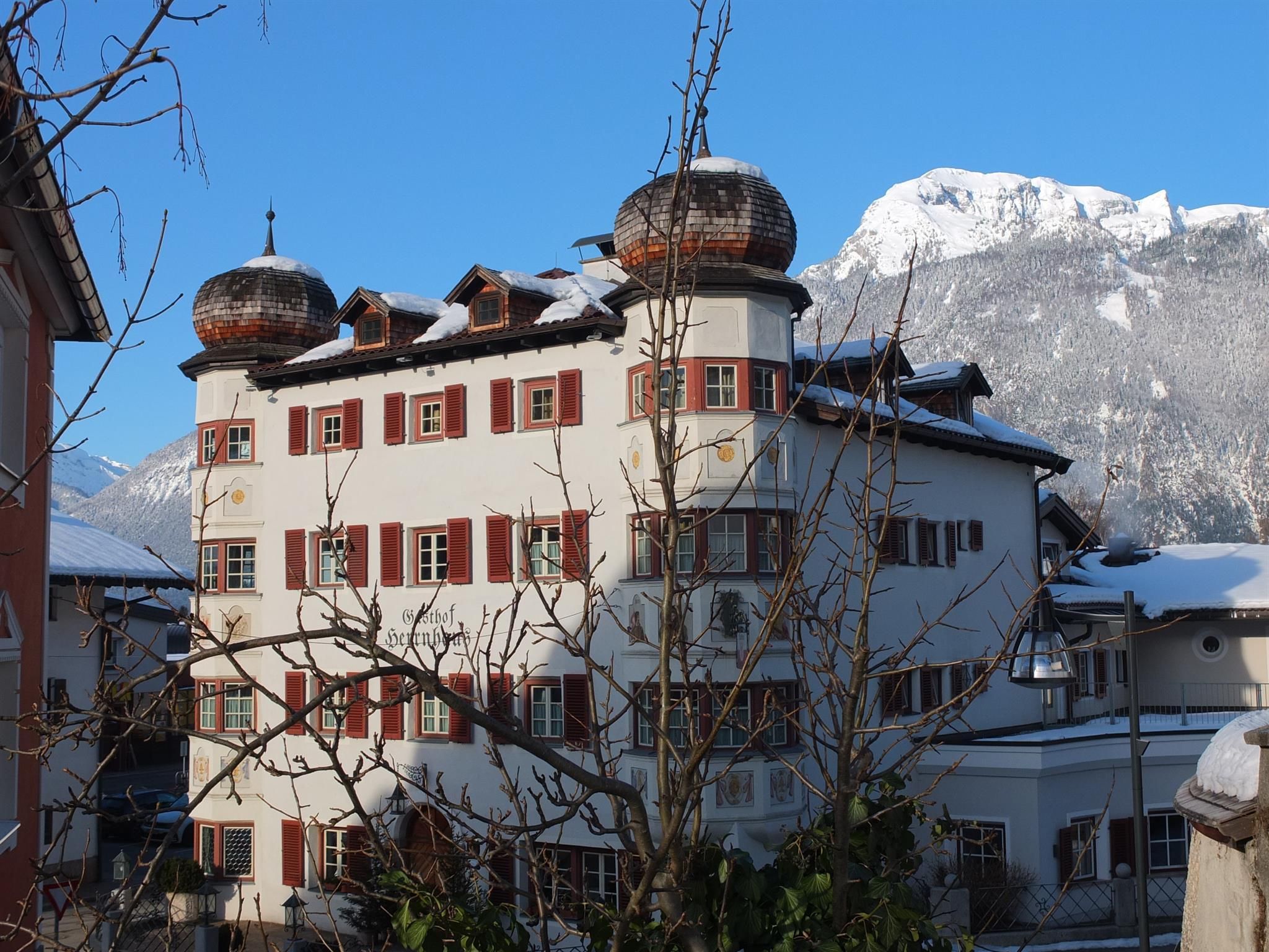 A historic building with round towers and red shutters. In the background, snow-covered mountains are visible under a clear blue sky.