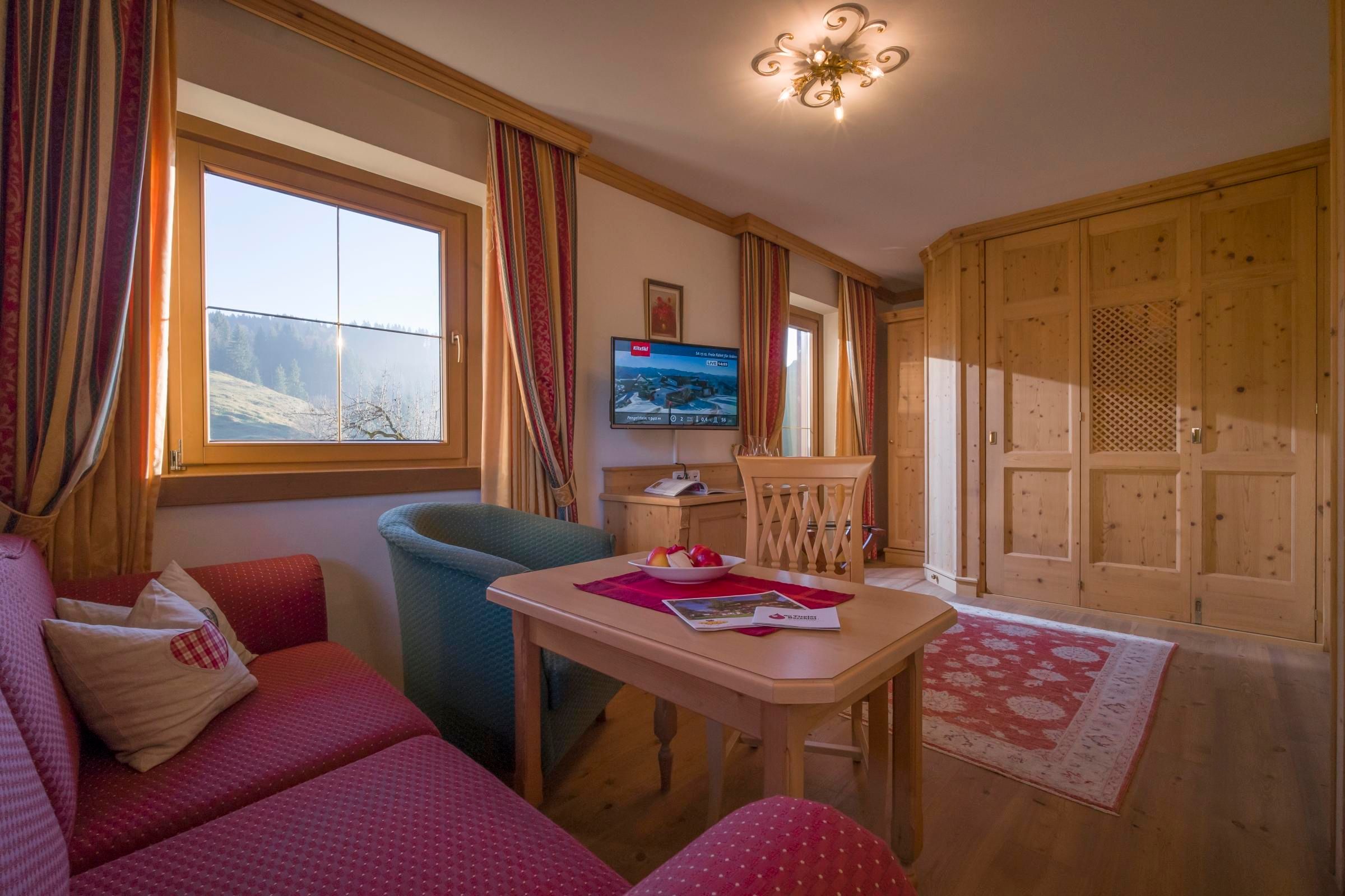 A cozy living room with a red sofa and a blue armchair. A wooden table and a television are also visible, as well as a window with a view of the mountains.