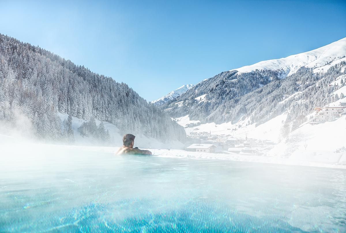 A man relaxes in a water basin amidst a snowy mountain landscape. The clear blue sky and the impressive mountains create a relaxing atmosphere.