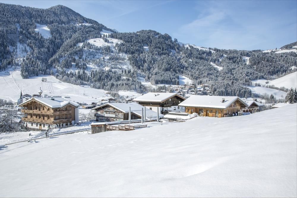A picturesque winter landscape with snowy hills and traditional chalets. The mountains in the background are also covered in snow and the sky is clear.