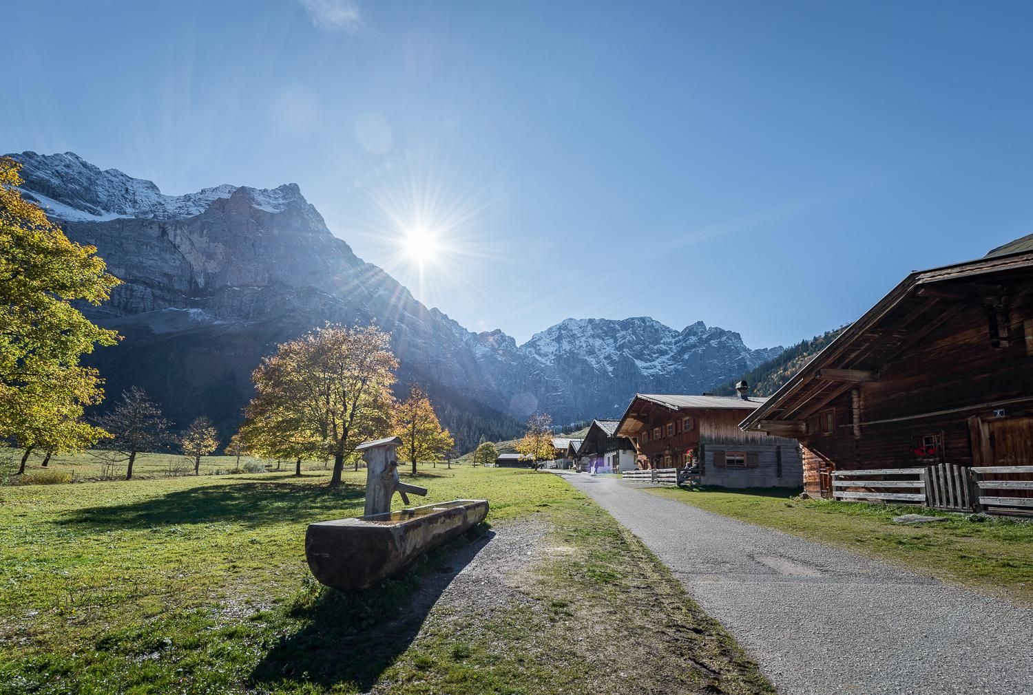 A picturesque landscape with tall mountains and bright sunshine. On the left side, there are trees and traditional wooden houses.