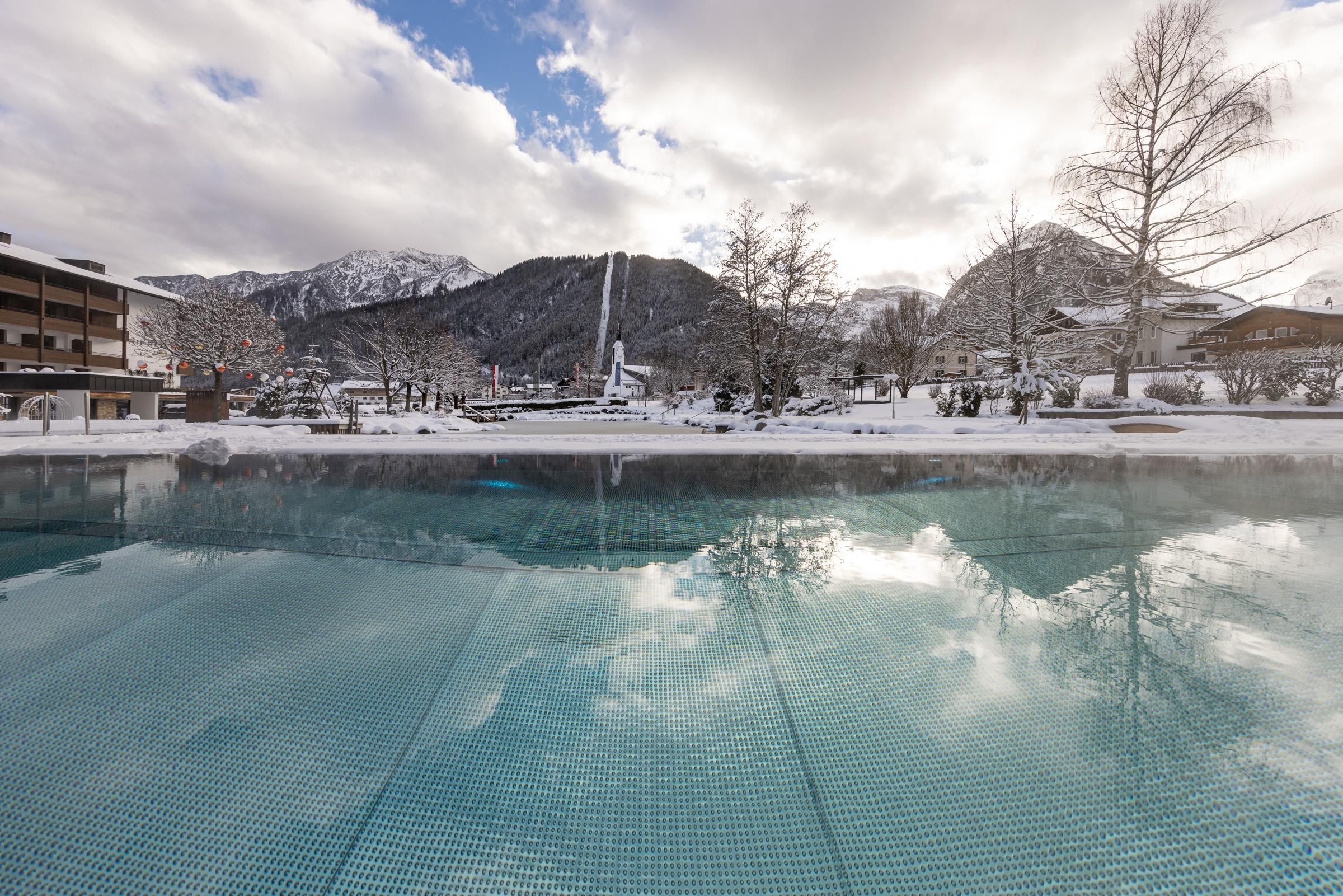 A winter view with a clear, reflecting pool and snow-covered mountains in the background. The clouds in the sky give the scene a calm atmosphere.