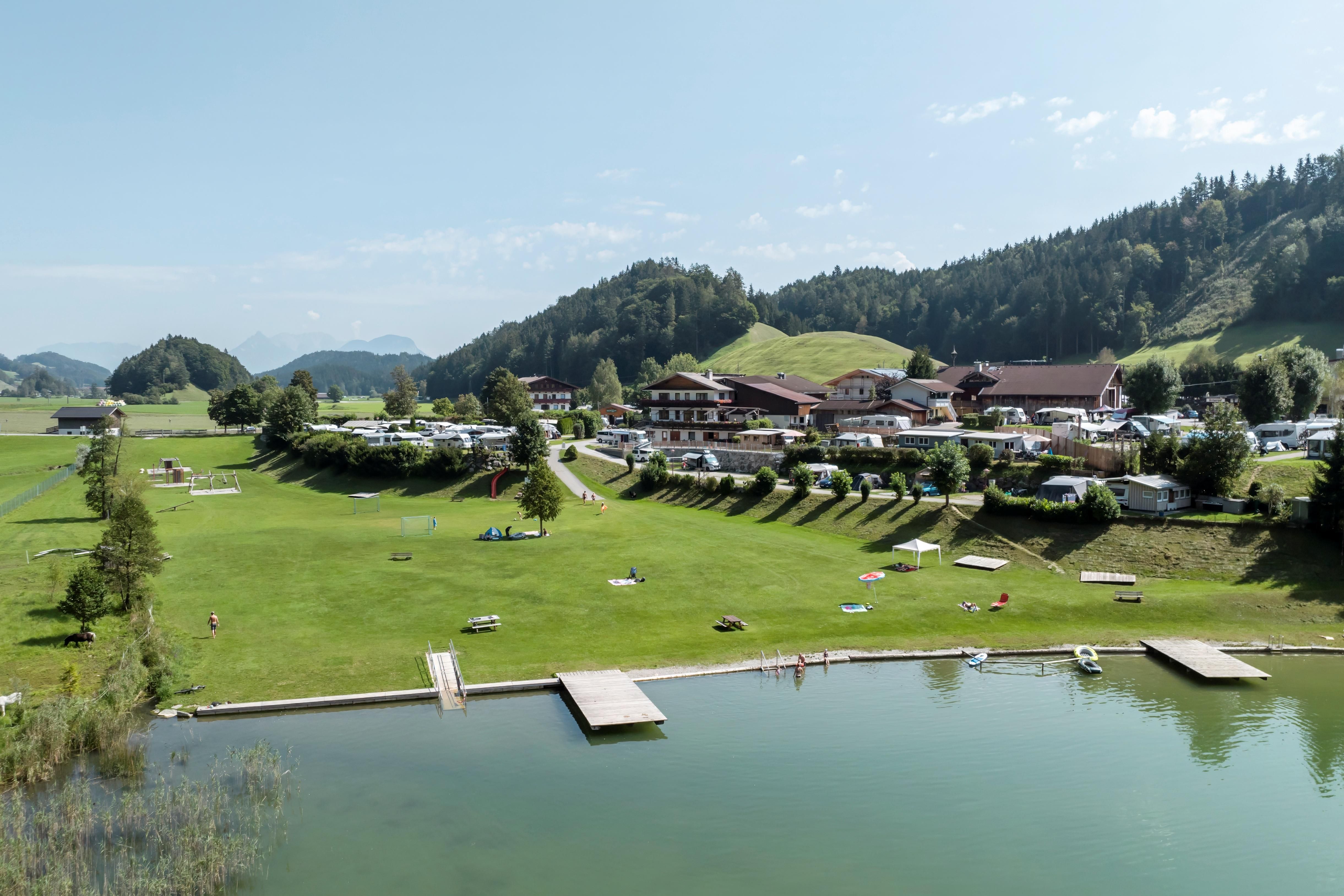 A beautiful landscape with meadows and a clear lake. In the background, you can see a cozy hotel and gentle hills.
