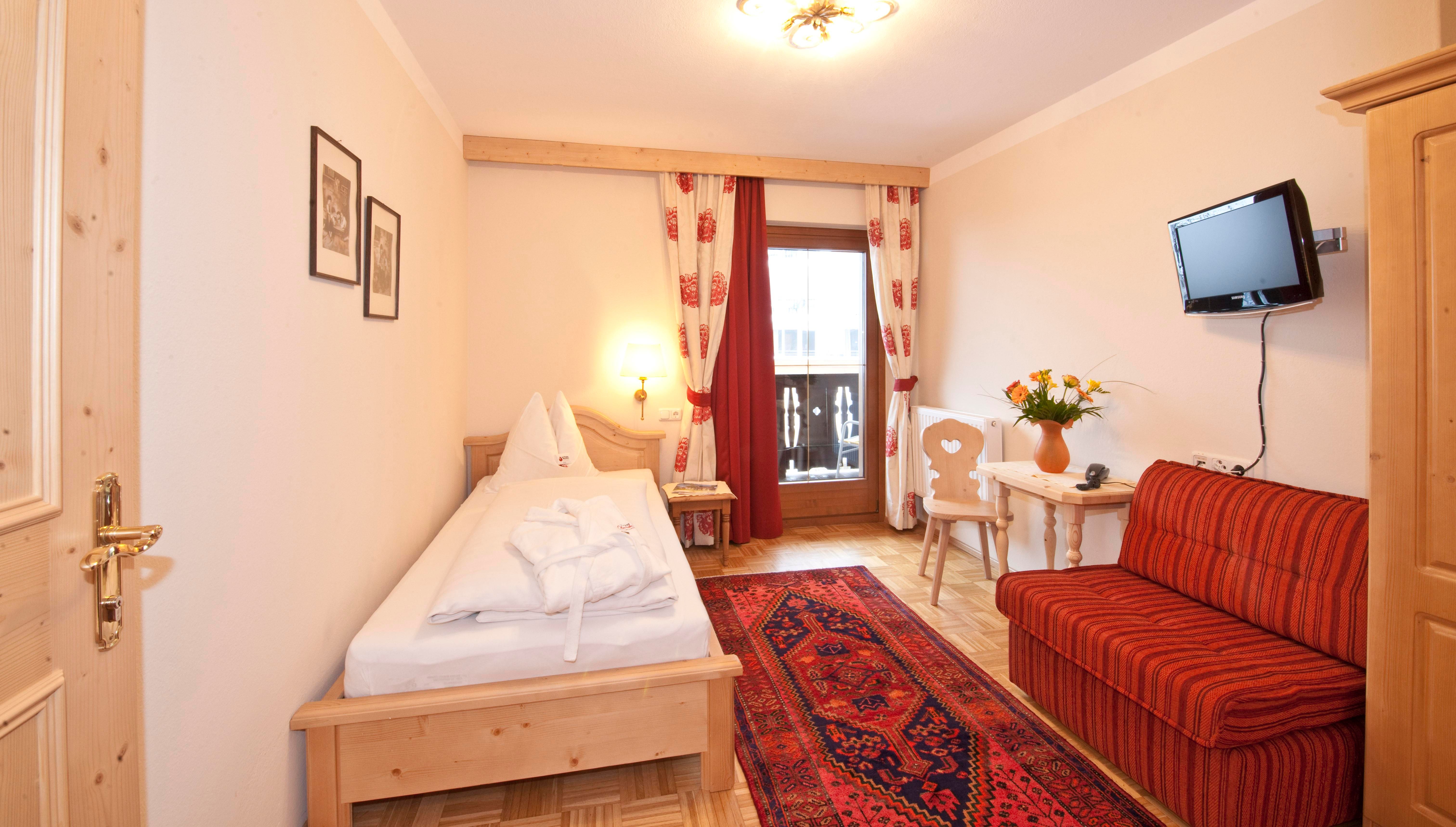 A cozy room with a single bed and a red sofa. A TV hangs on the wall, and there is a small table with flowers.