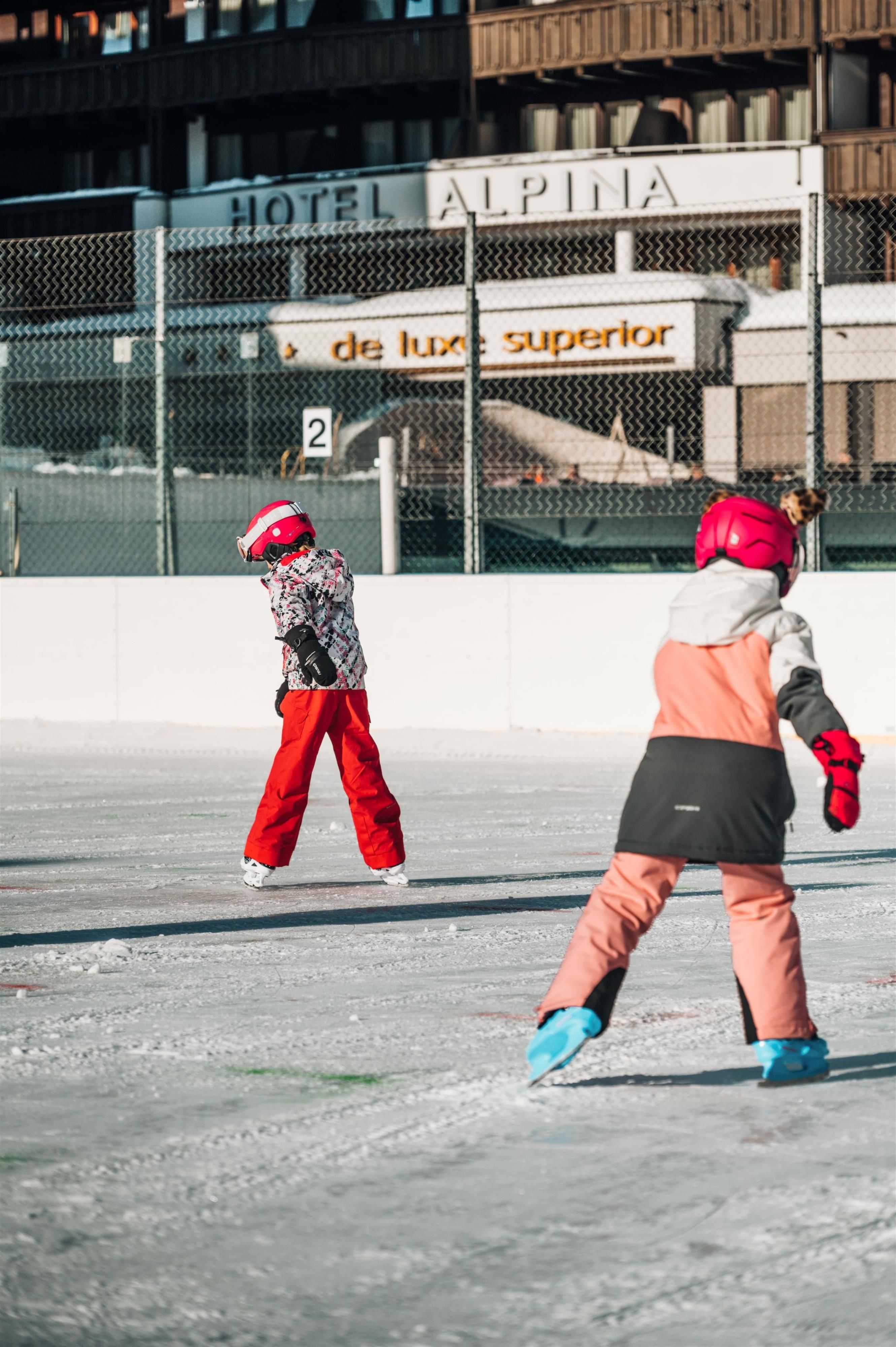 Two children are ice skating on an ice rink. In the background, the Hotel Alpina is visible.