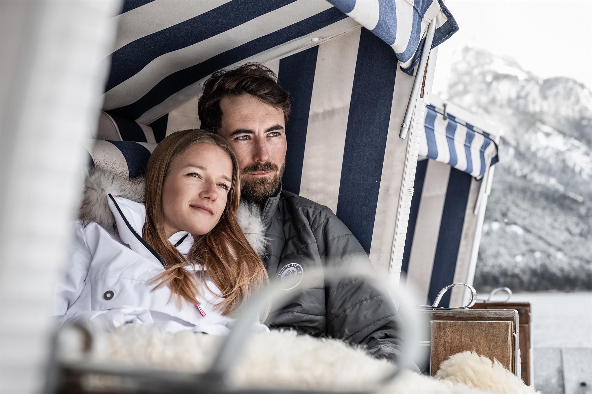 A couple is comfortably sitting in a beach chair and enjoying the view of the winter landscape. They are warmly dressed and seem relaxed and happy.